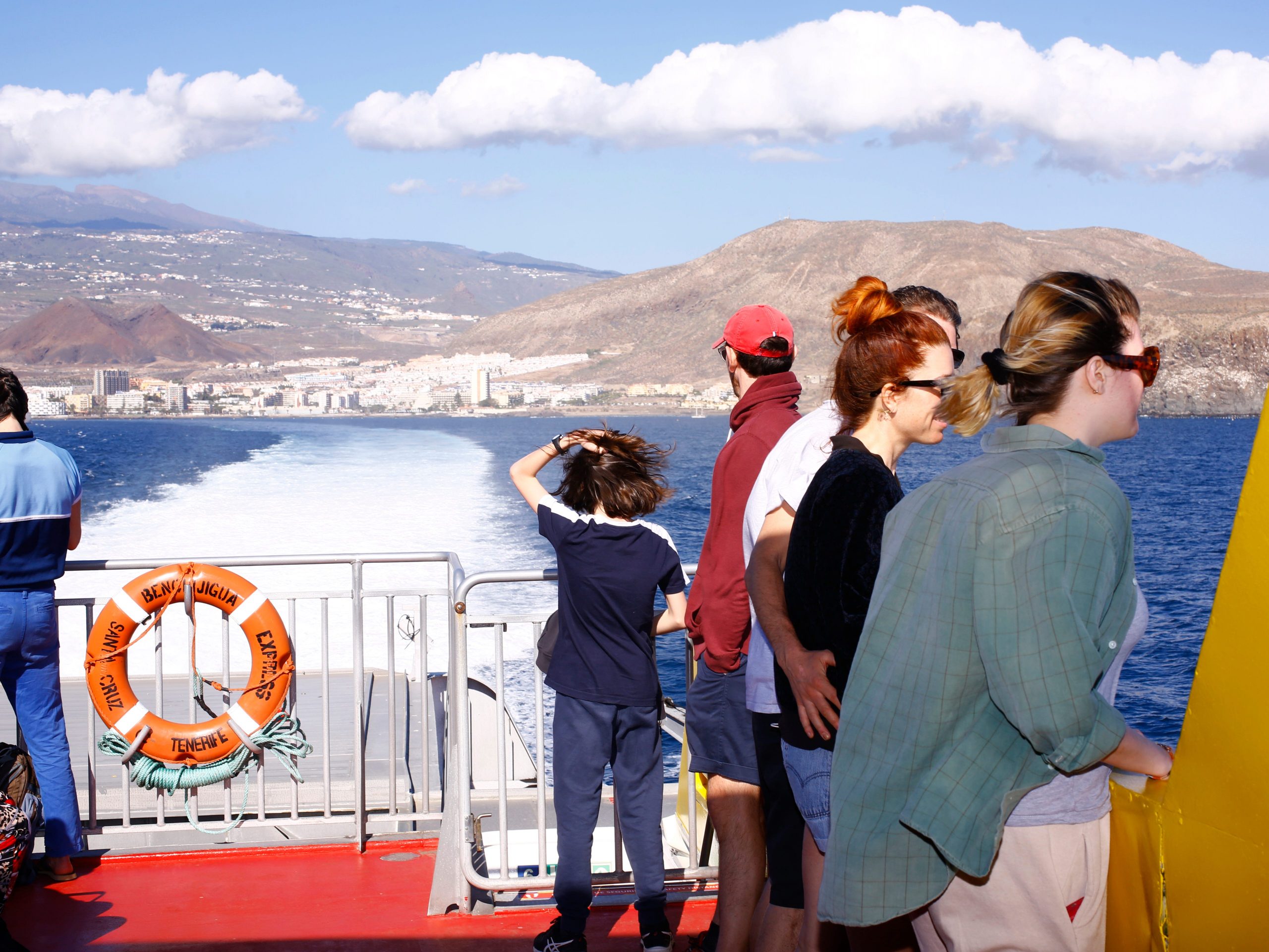 Several people standing inside a boat moving in the ocean with a city surrounded by mountains in the background.