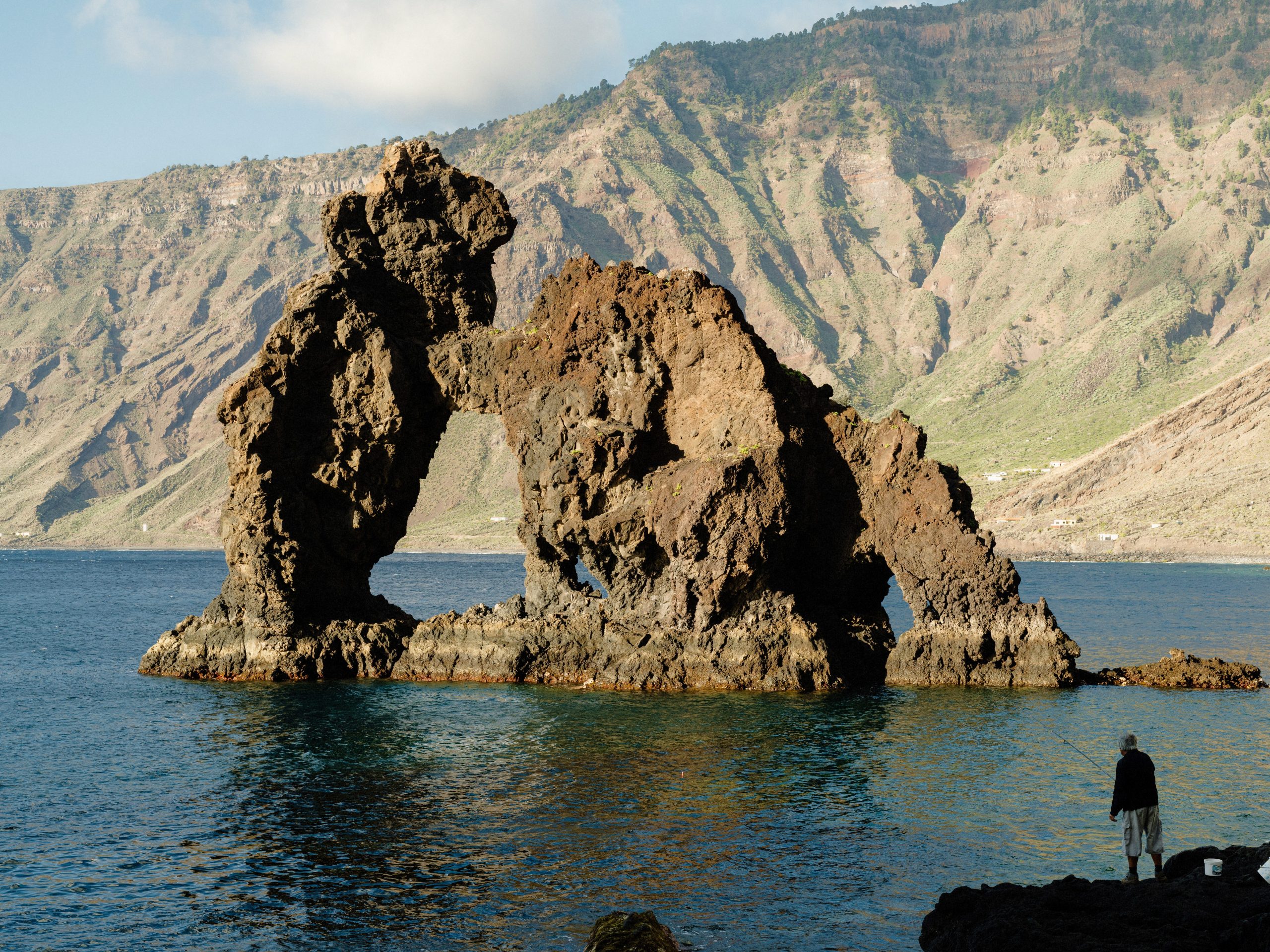 A person standing in front of a large rock formation rising out of the water in front of a mounntain.