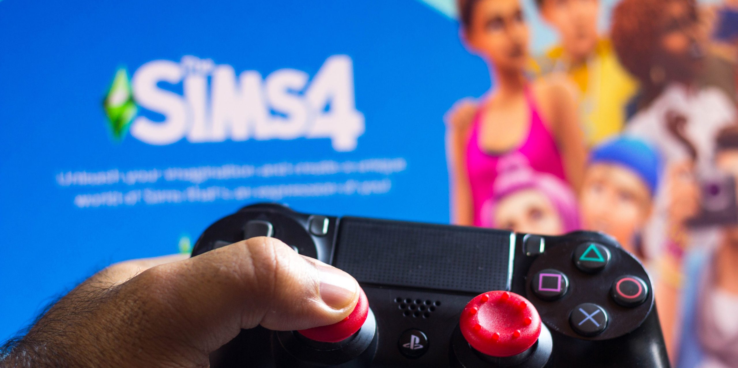A person's hand holding a video game controller in front of a screen showing The Sims 4.