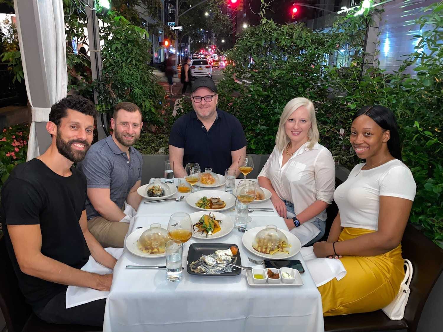 SevenRooms employees, three men and two women, sit around a table at a restaurant. There is lots of food on the table, which is surrounded by plants.