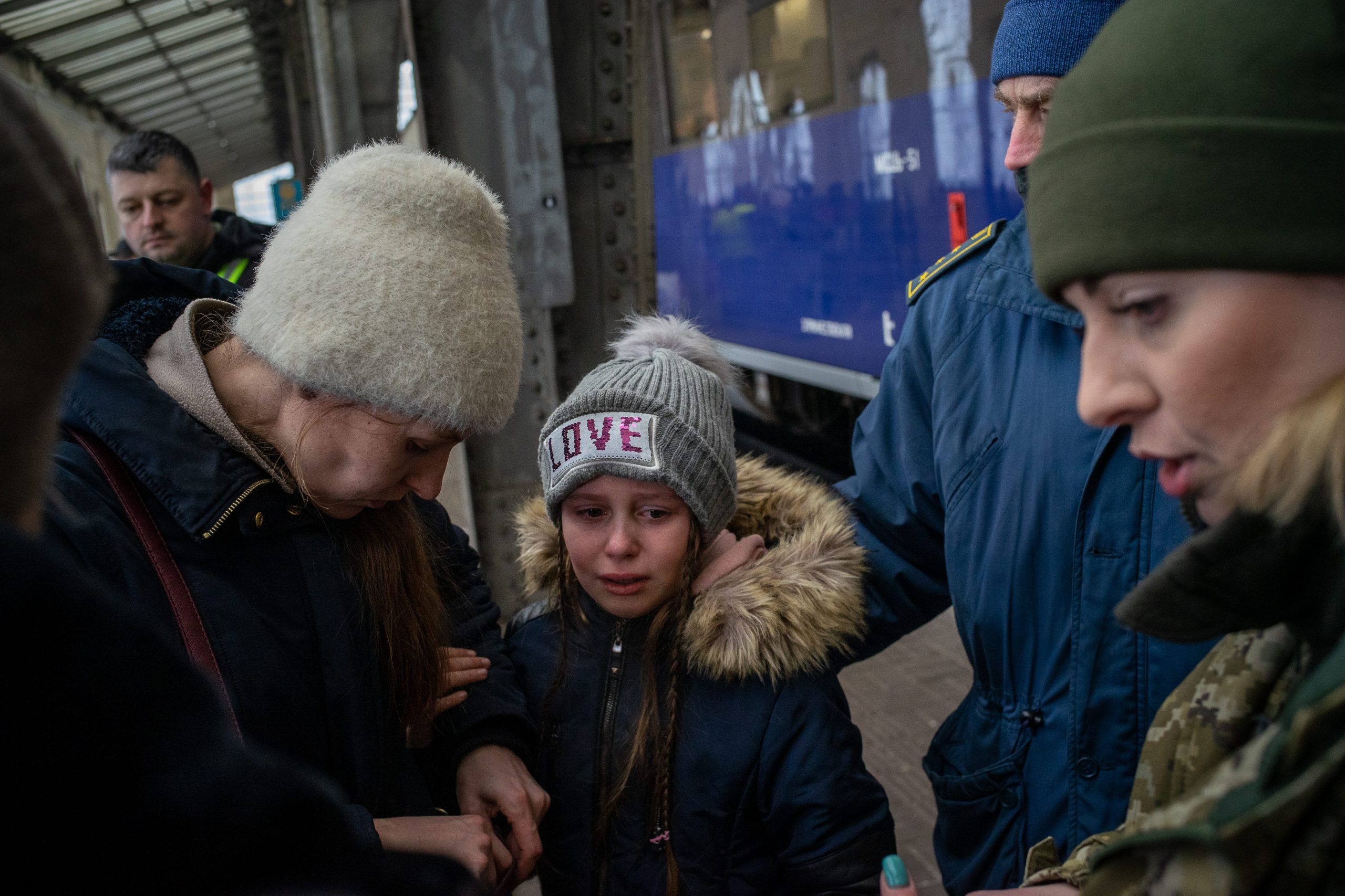 A girl with a hat that reads "Love" is surrounded by several adults and cries as she stands on a train platform.