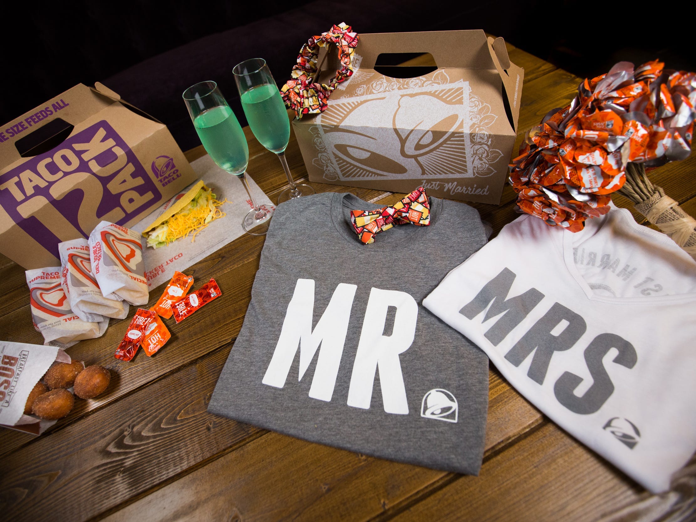 Taco Bell food boxes, Mr. and Mrs. shirts, hot sauce packet bow tie and flower bouquet, green drinks in champagne glasses, and some food.