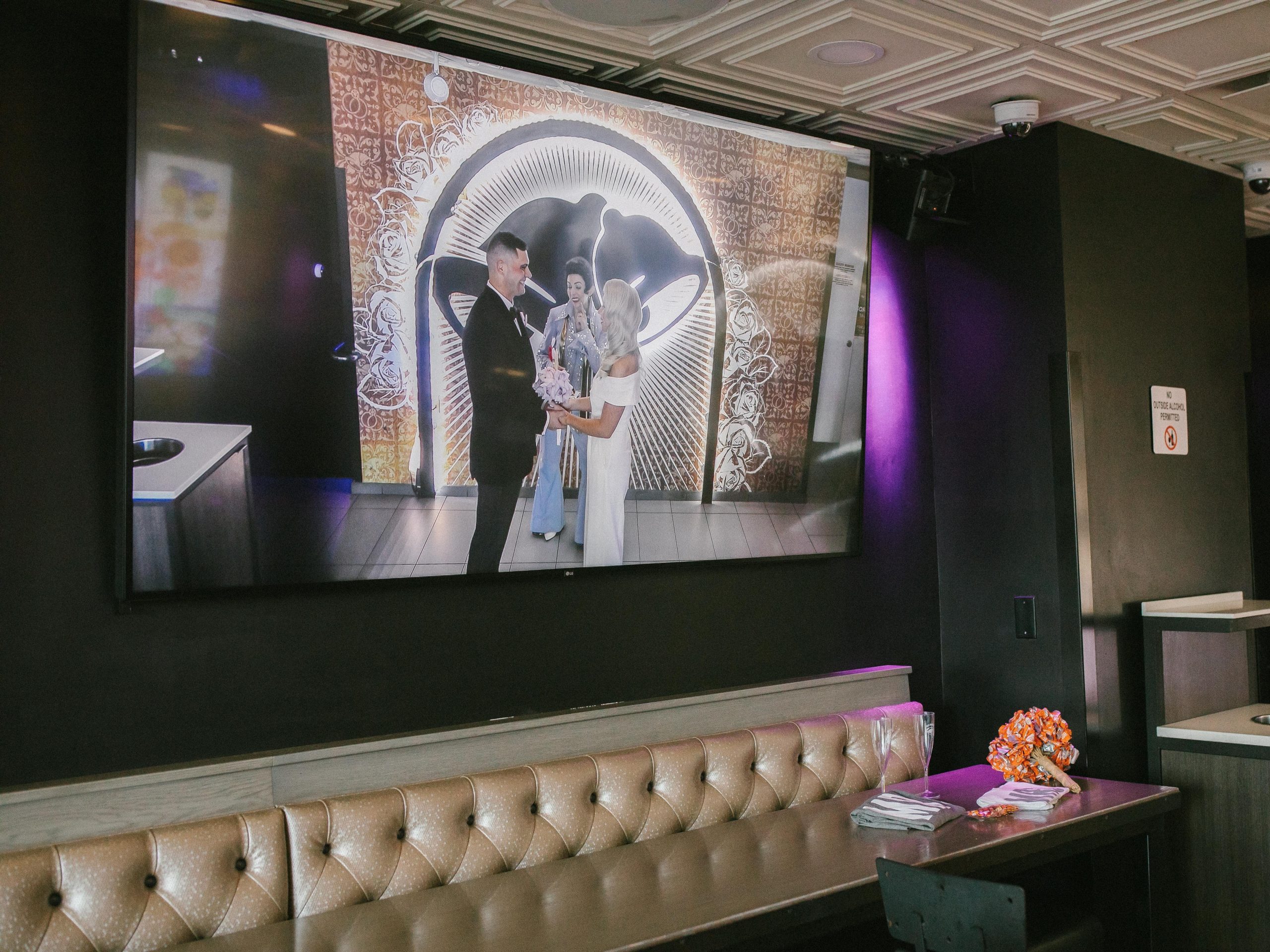 Side of chapel with large tv screen showing a couple standing in front of the bell archway. It hangs over a long booth and table against the wall.