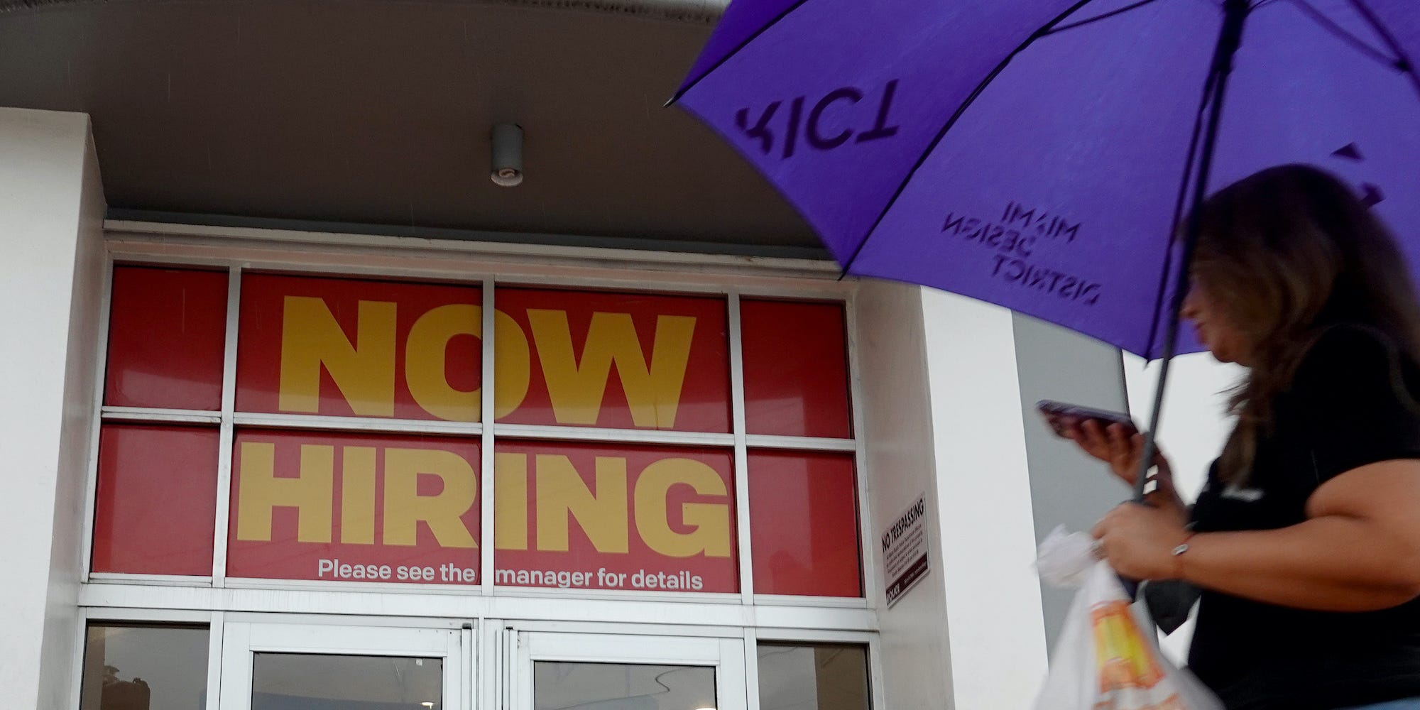 A person with an umbrella walks by a McDonald's restaurant that has a ''Now Hiring" sign above the entrance.