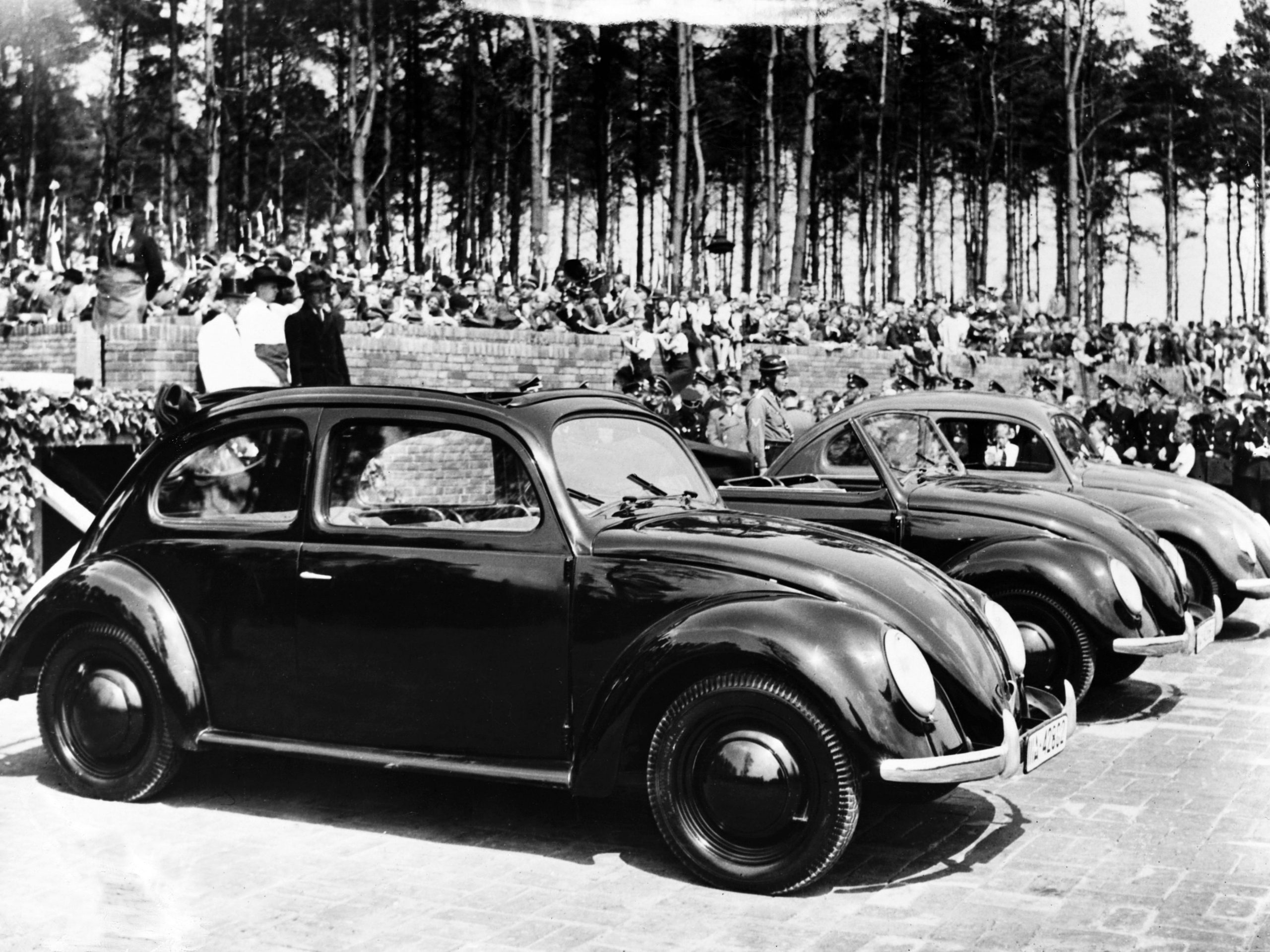 Adolf Hitler staat achter de auto's in een donkere jas, klaar om de nieuwe fabriek in te wijden.