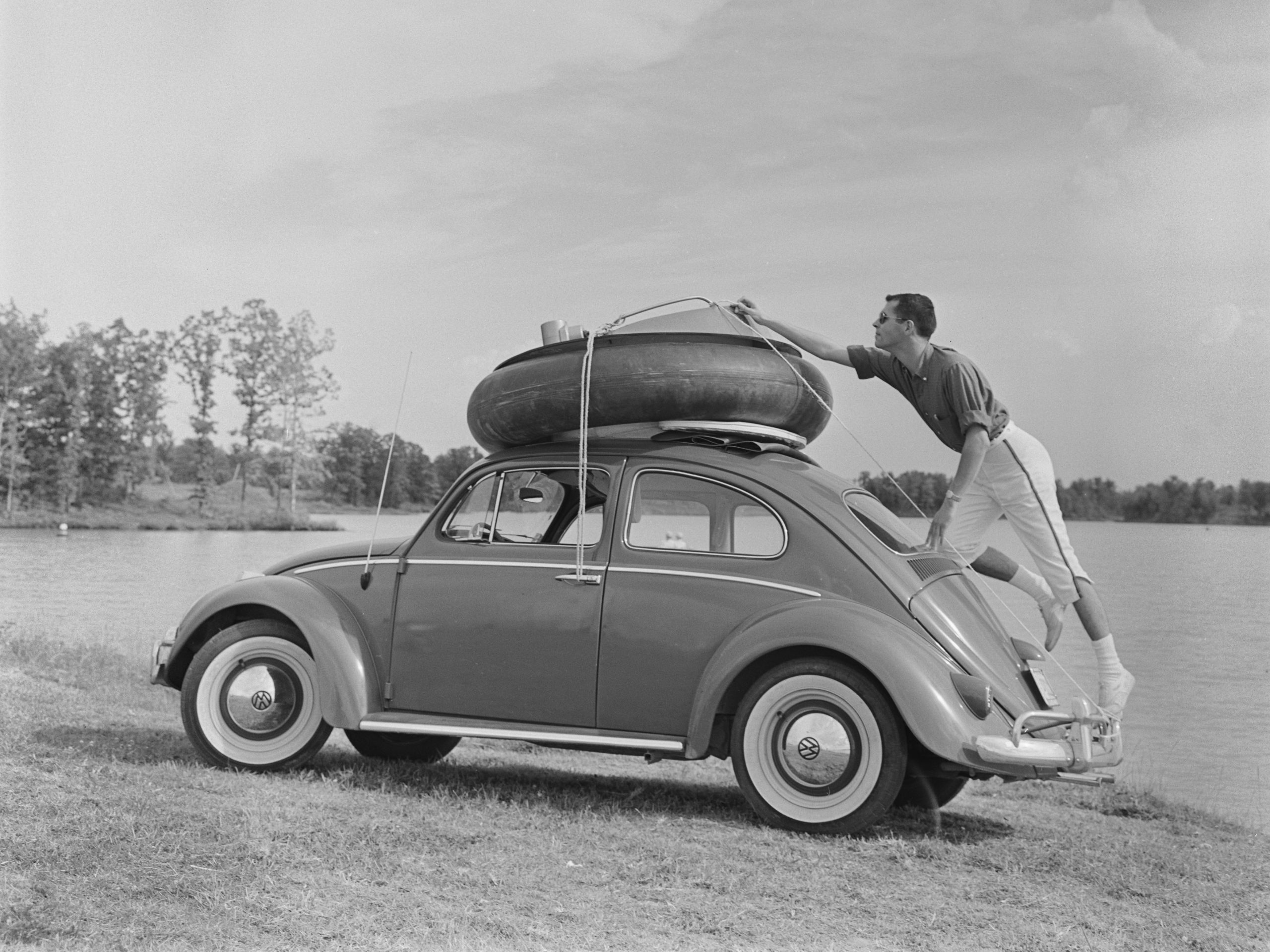 man staat op de achterbumper van een Volkswagen Kever terwijl hij de touwen aanpast die een binnenband en een rubberbootadapter aan het dak van de auto binden, locatie onbekend, Verenigde Staten, circa 1955.