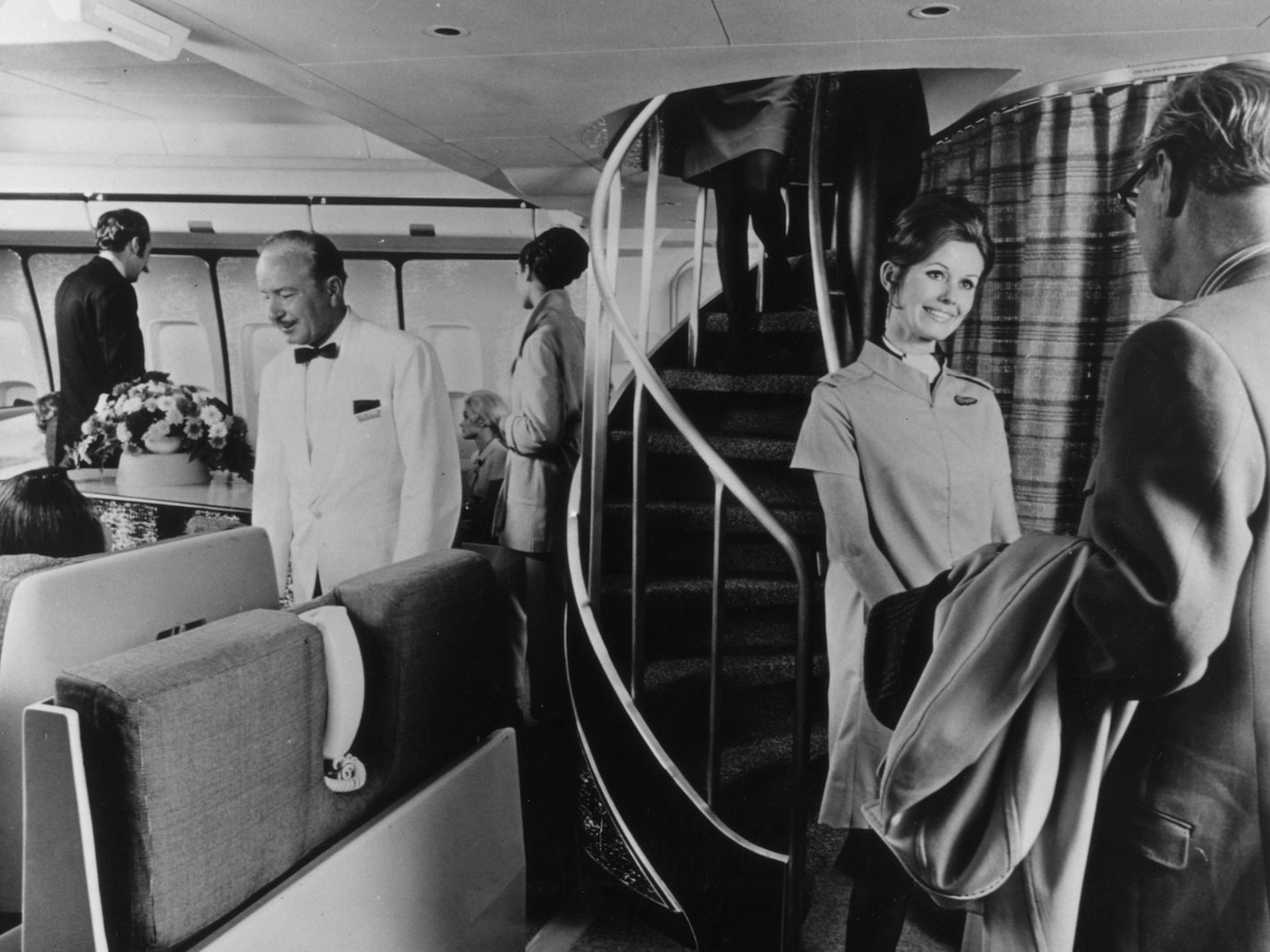 A BOAC air hostess greets a passenger in front of a spiral staircase which leads to the upper deck lounge in a Boeing 747 Monarch.