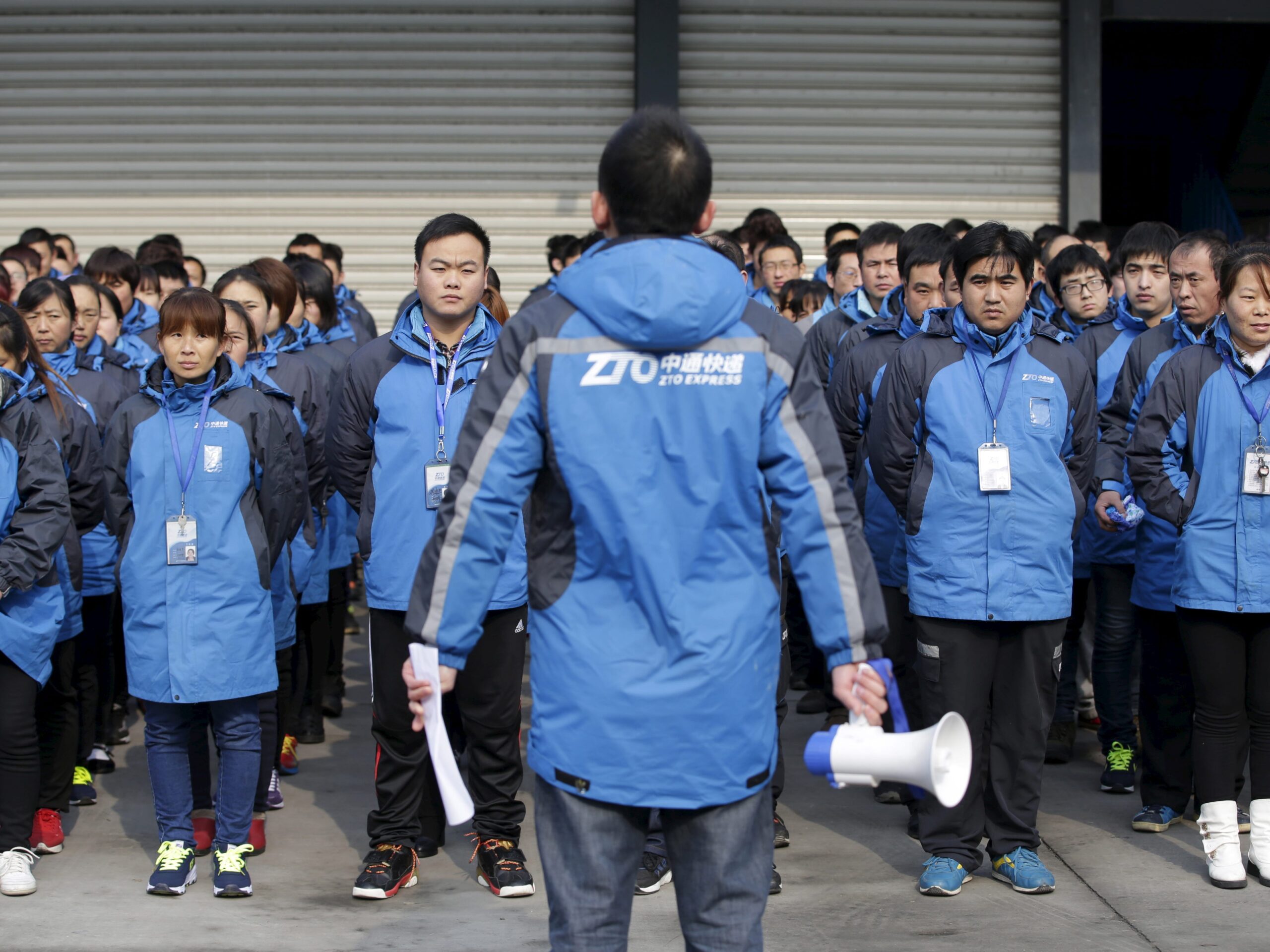 Chinese people stand in straight lines wearing blue jackets that say ZTO Express as one person addresses the group.