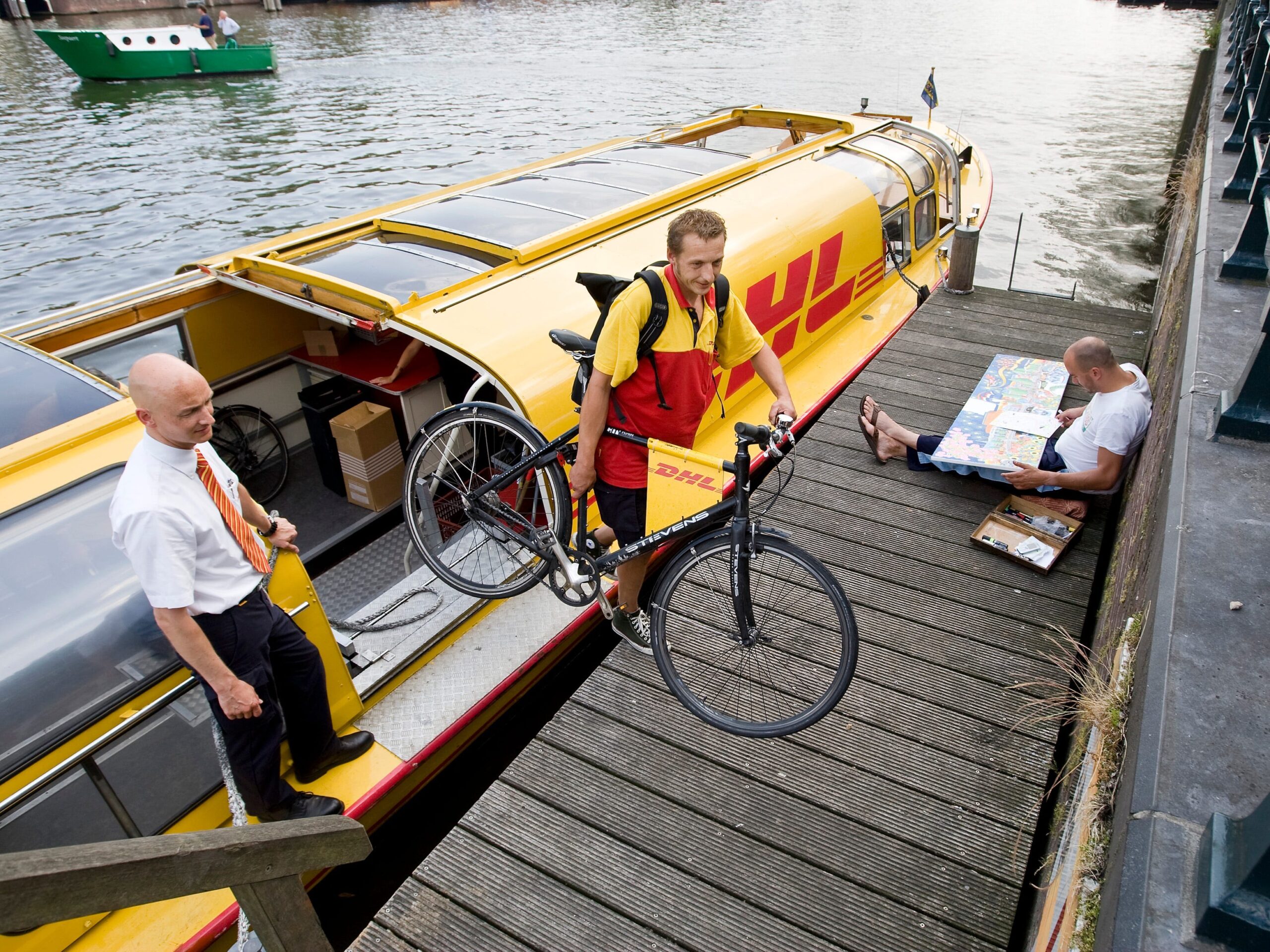 A man in a red and yellow DHL uniform lifts a bicycle off of a yellow, DHL-branded river barge while the boat driver observes.