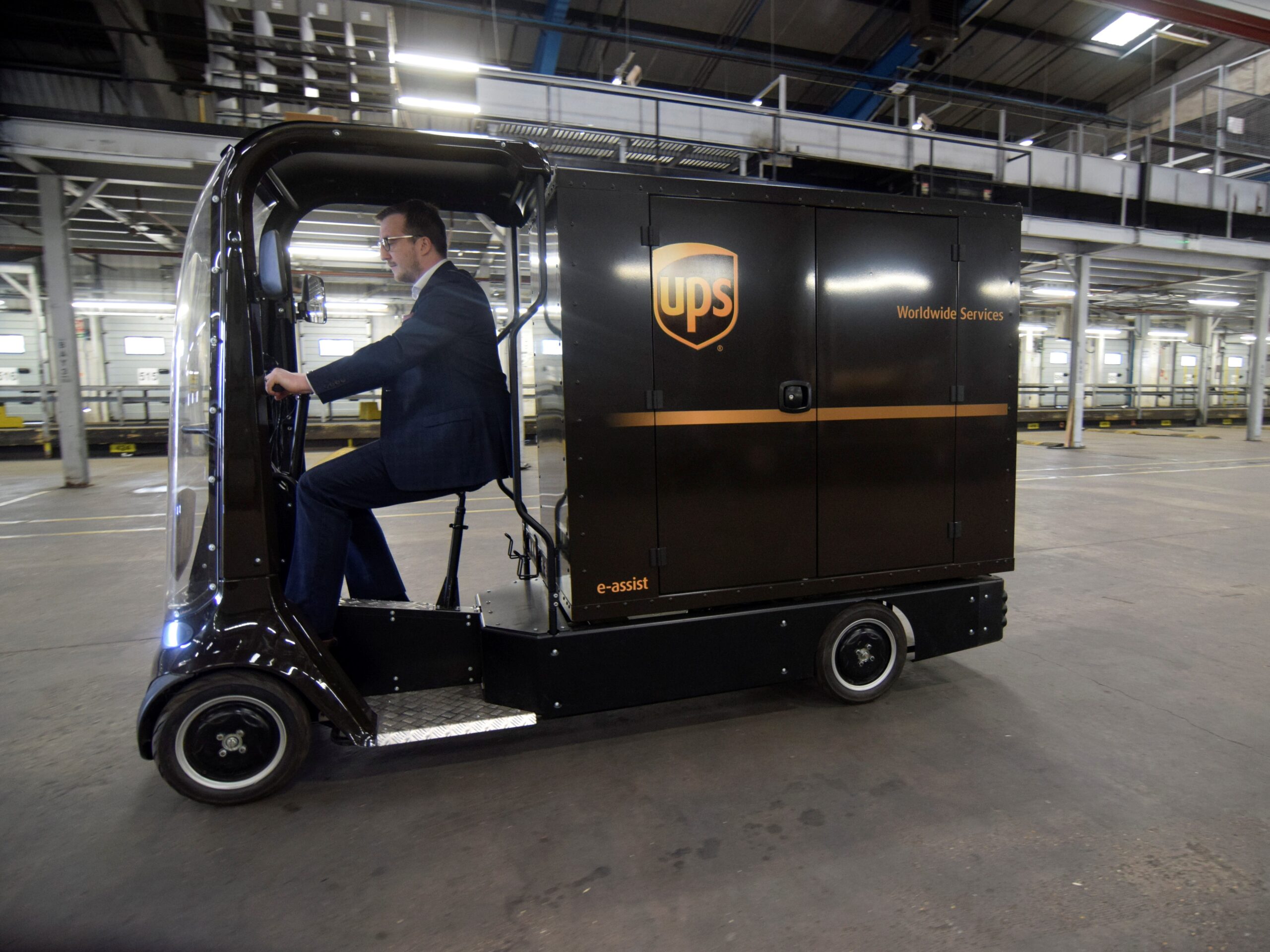 A white man in a suit drive a 4-wheeled brown cycle with the UPS logo on the side.