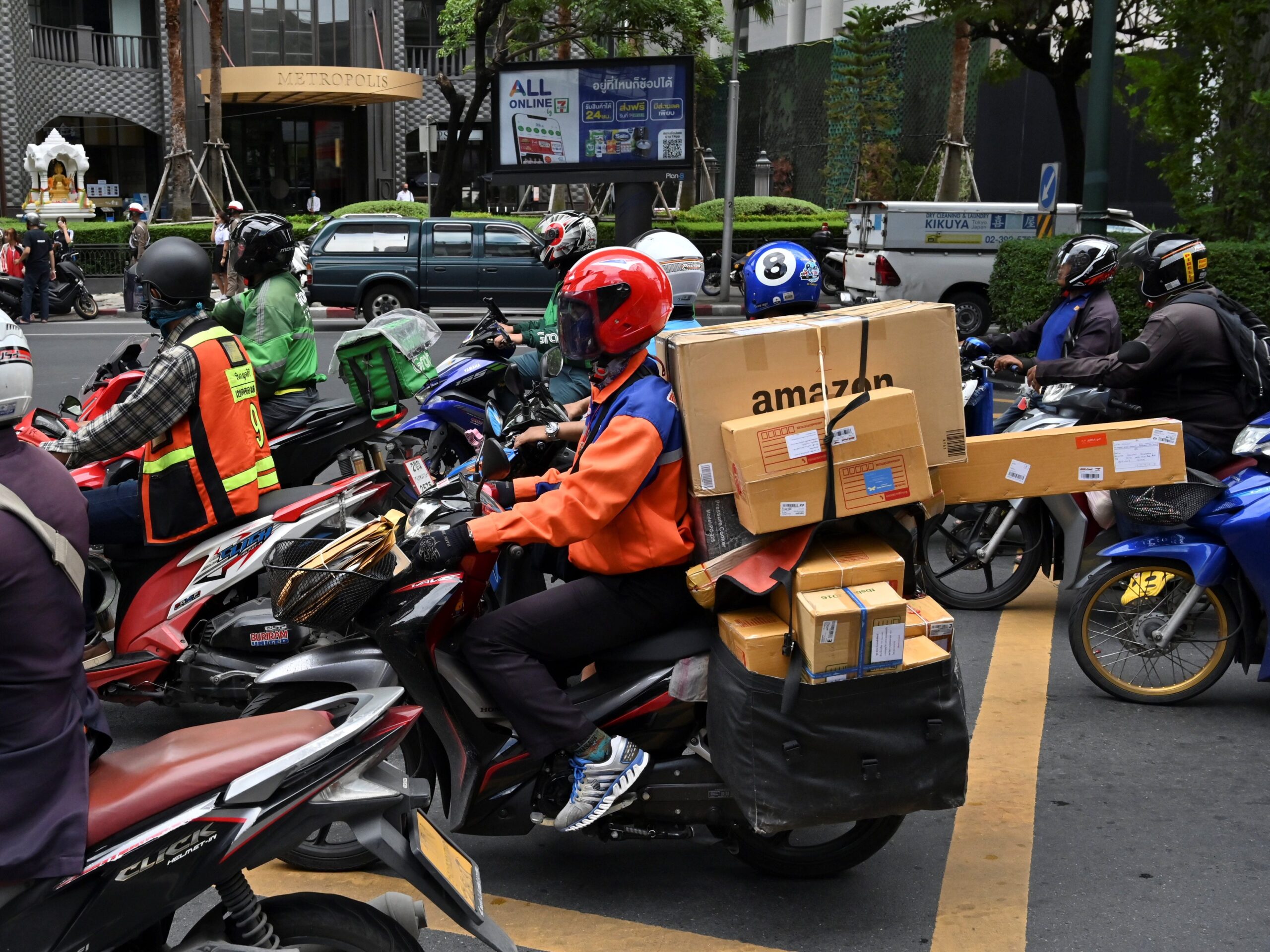 A man in a orange jacket and red helmet rides a motor bike. The back is stuffed with cardboard packages and one has the Amazon logo.