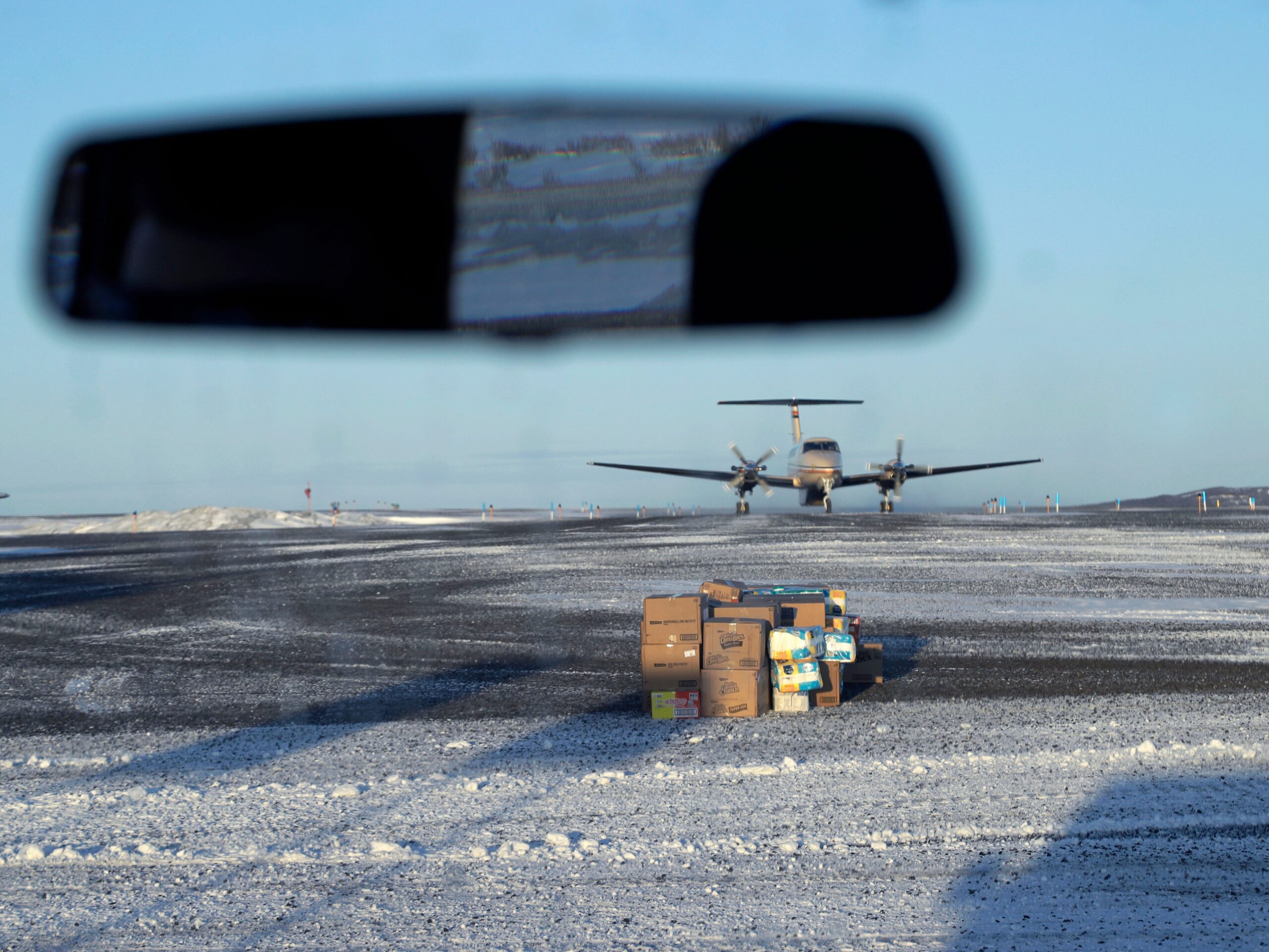 A pile of boxes sits on a snowy tarmac as with a plane in the background.
