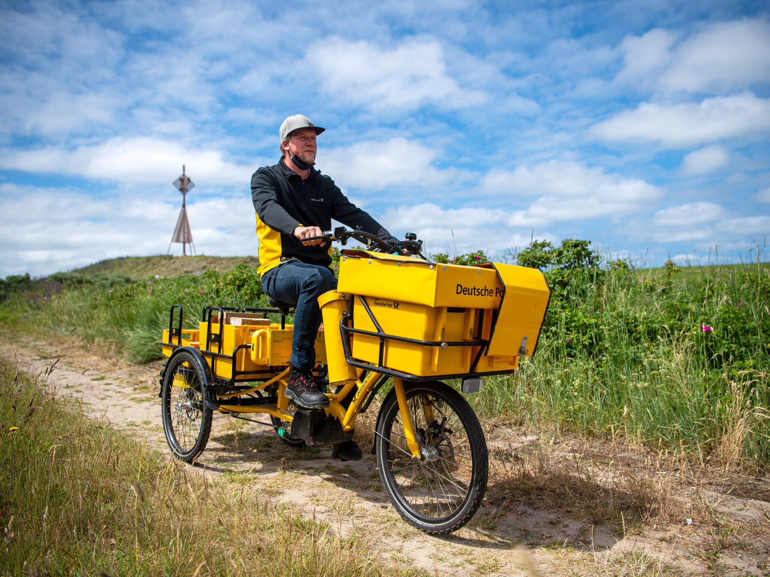 A man in jeans and a black and yellow short ride a tricycle with large cargo baskets on the front and back through a field.