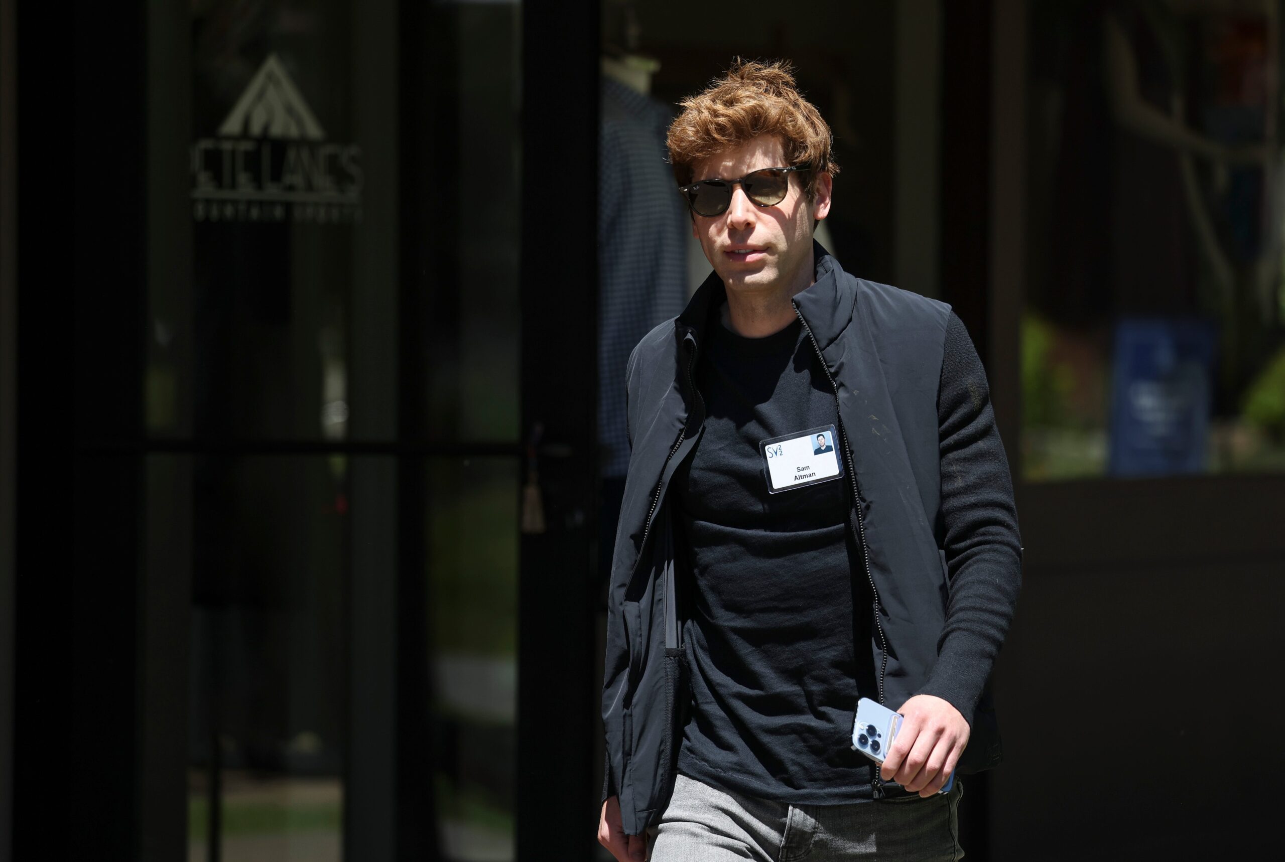 sam altman wearing a black t shirt, black jacket, grey pants and sunglasses