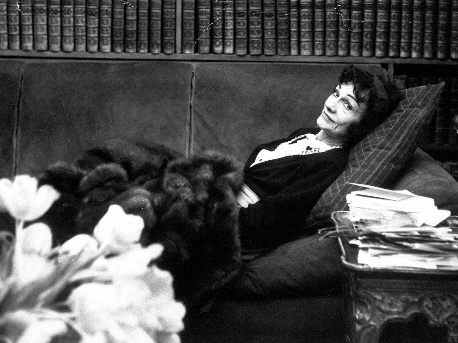 Coco Chanel lounging on a couch in front of bookshelves