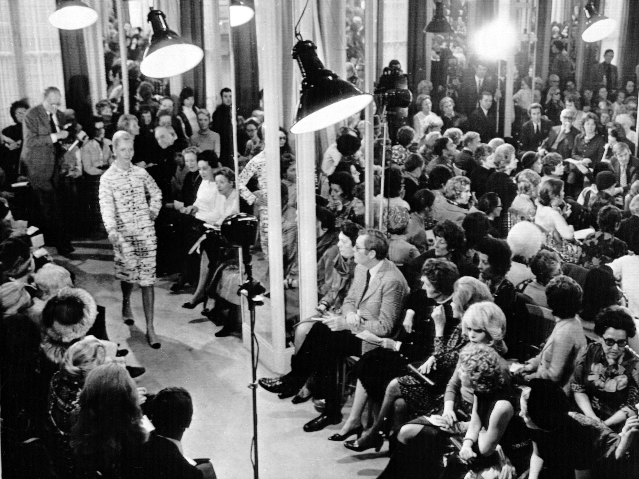 People sit in crowded space watching model wearing Chanel walk down runway