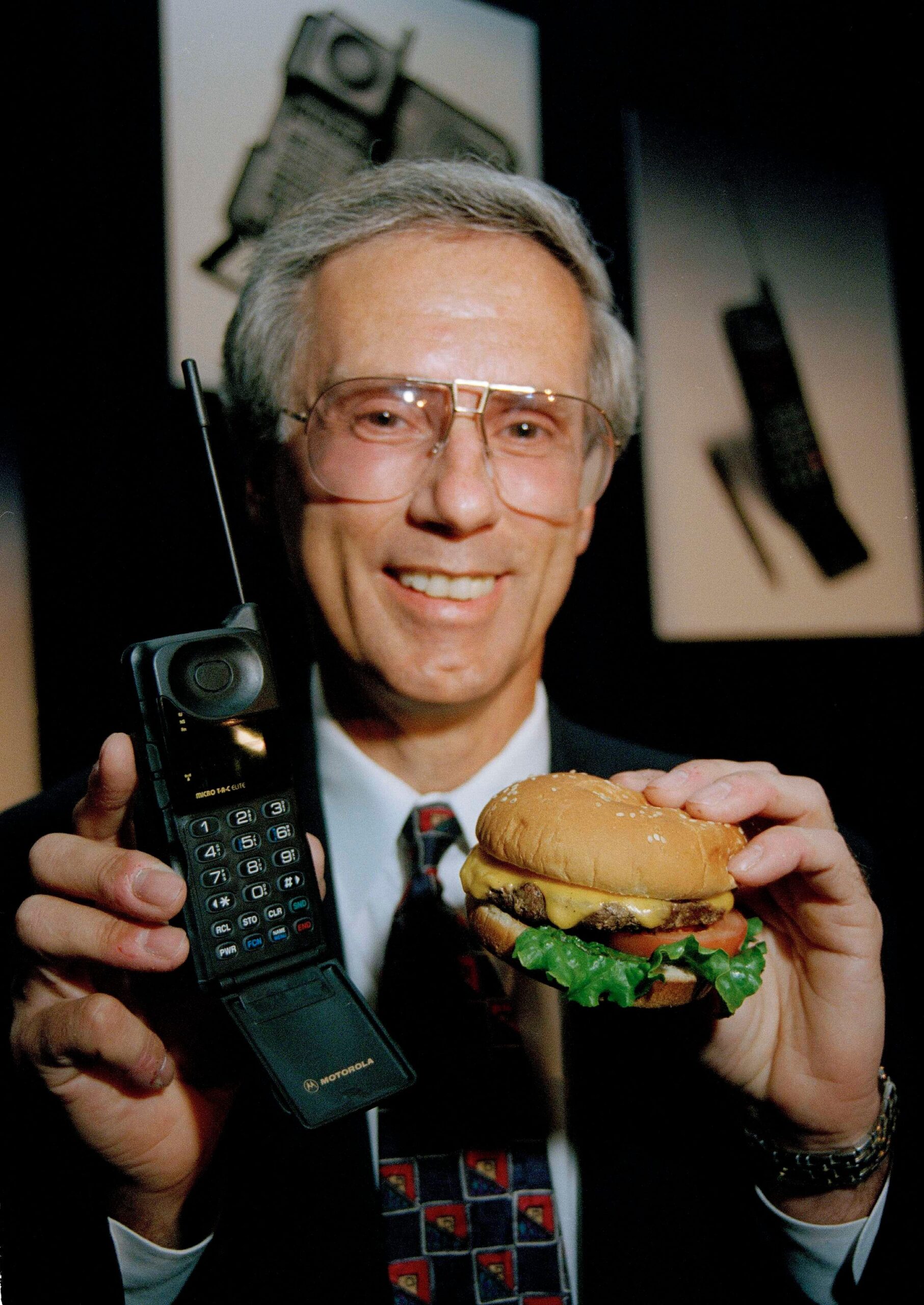 man met grijs haar in een pak met in zijn ene hand een Motorola MicroTac Elite mobiele telefoon en in zijn andere hand een hamburger om de maten te vergelijken.