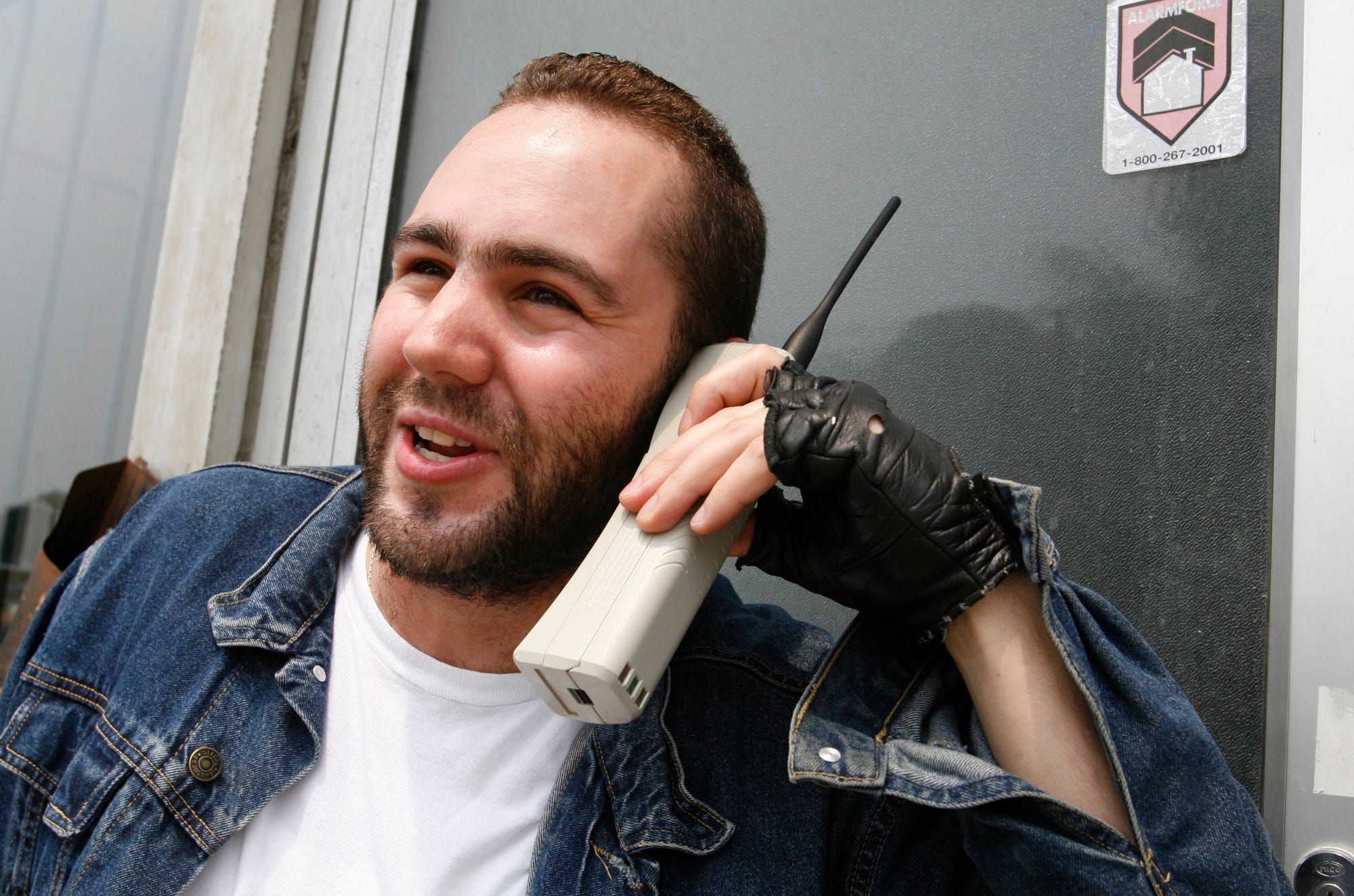 een man met een wit T-shirt, spijkerjack en zwarte vingerloze handschoen, praat met een witte stenen telefoon...