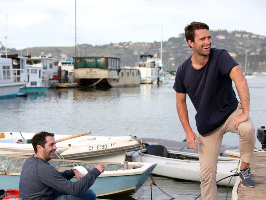 Joey Zwillinger, who is sitting in a boat, and Tim Brown posing for a picture on a dock with boats moored around them.