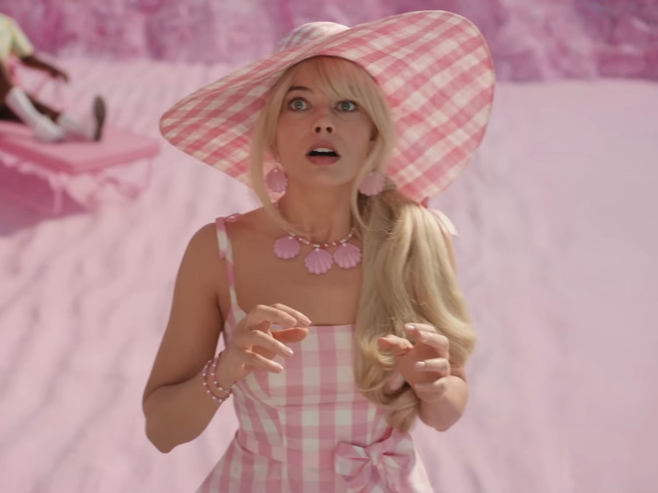 Margot Robbie in a scene from &#34;Barbie&#34; where Barbie is standing on a beach and looking up with a shocked expression on her face while wearing a pink and white plaid dress with a large sunhat.