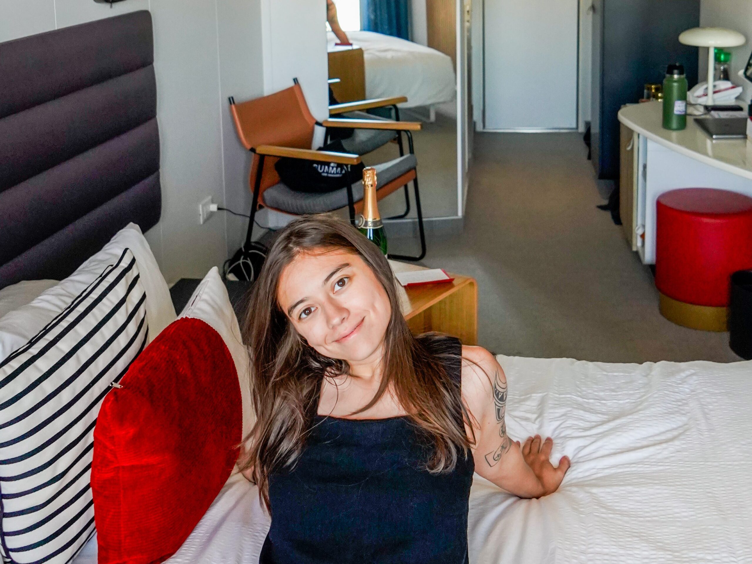 Left: The author in a white tank and sunglasses in front of boats on a dock with a cruise ship in the background. Right: The author sits on a bed with white sheets and a red pillow on the left. Behind her is the cruise ship cabin