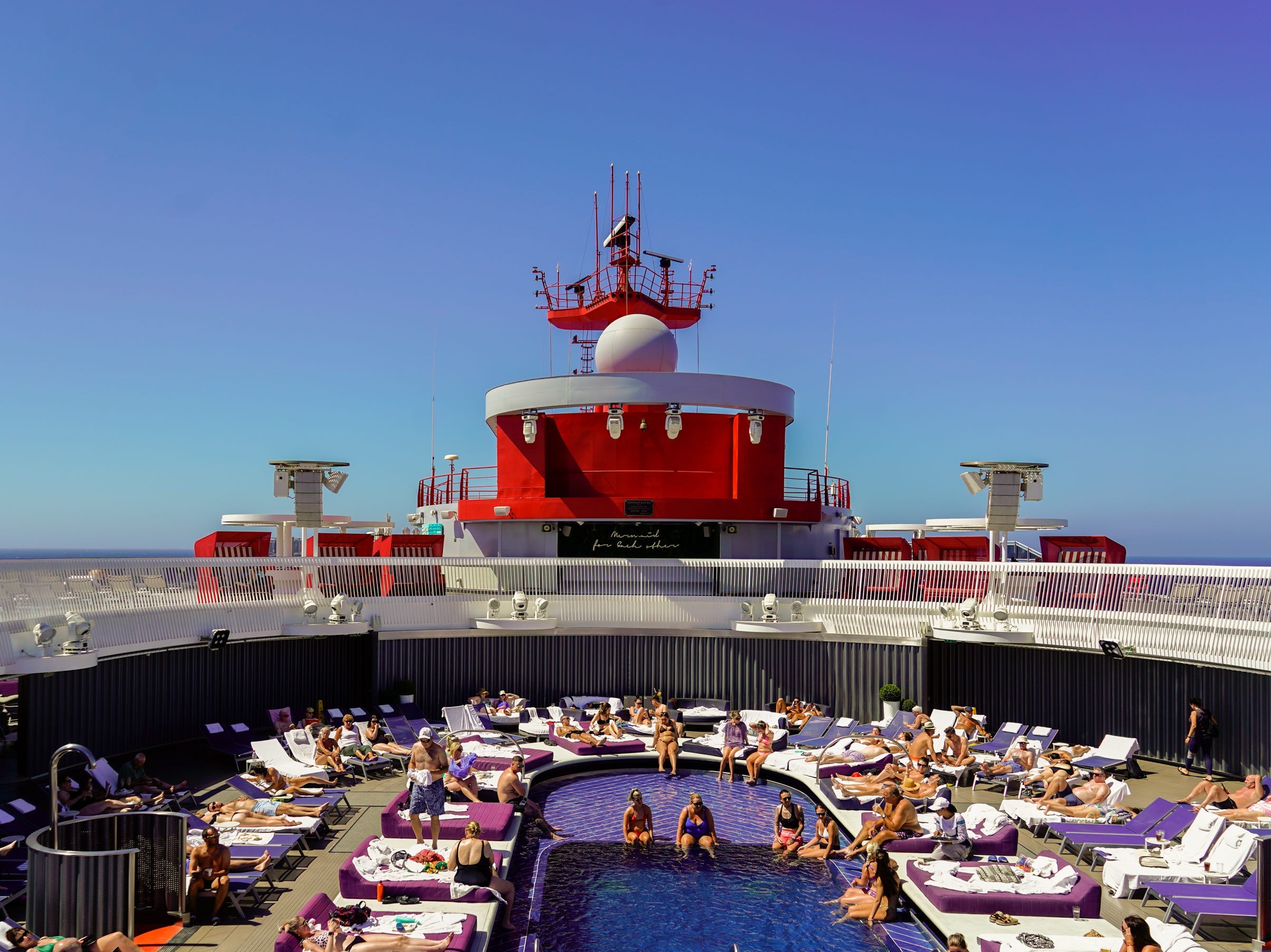 A pool on a cruise ship with people inside it and on pool chairs around it. The sky is clear and blue behind the ship.