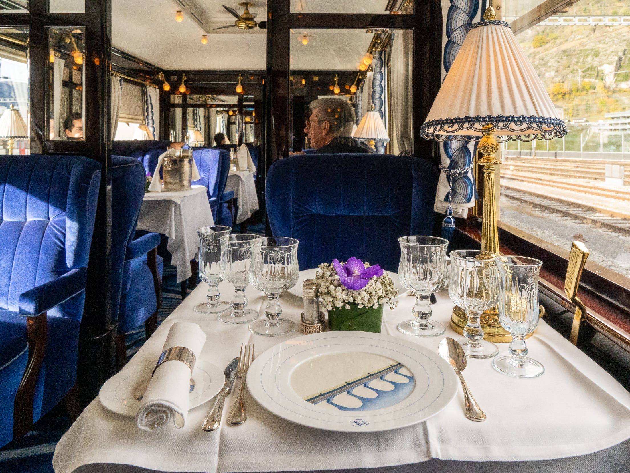 Inside a train dining car with velvet, blue seats, and tables with white cloths, dishes, and silverware