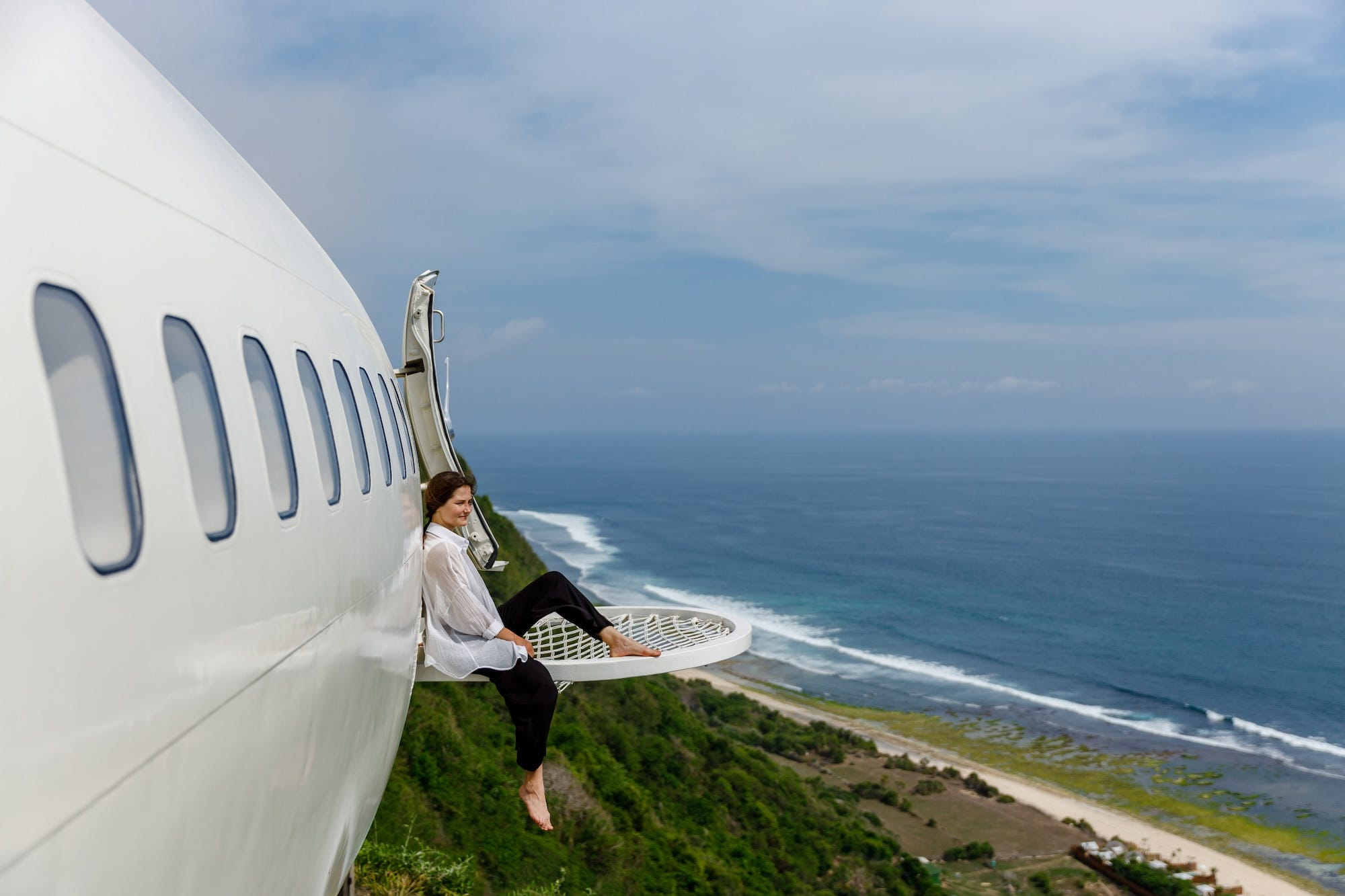 Een vrouw zit in een hangmat aan de zijkant van een wit vliegtuig met uitzicht op een strand.