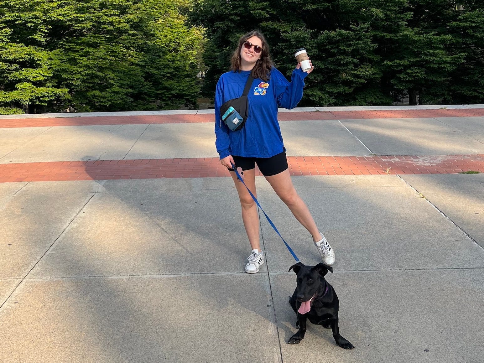 A woman standing with her dog, holding coffee and wearing a fanny pack. There are trees and a blue sky behind them.