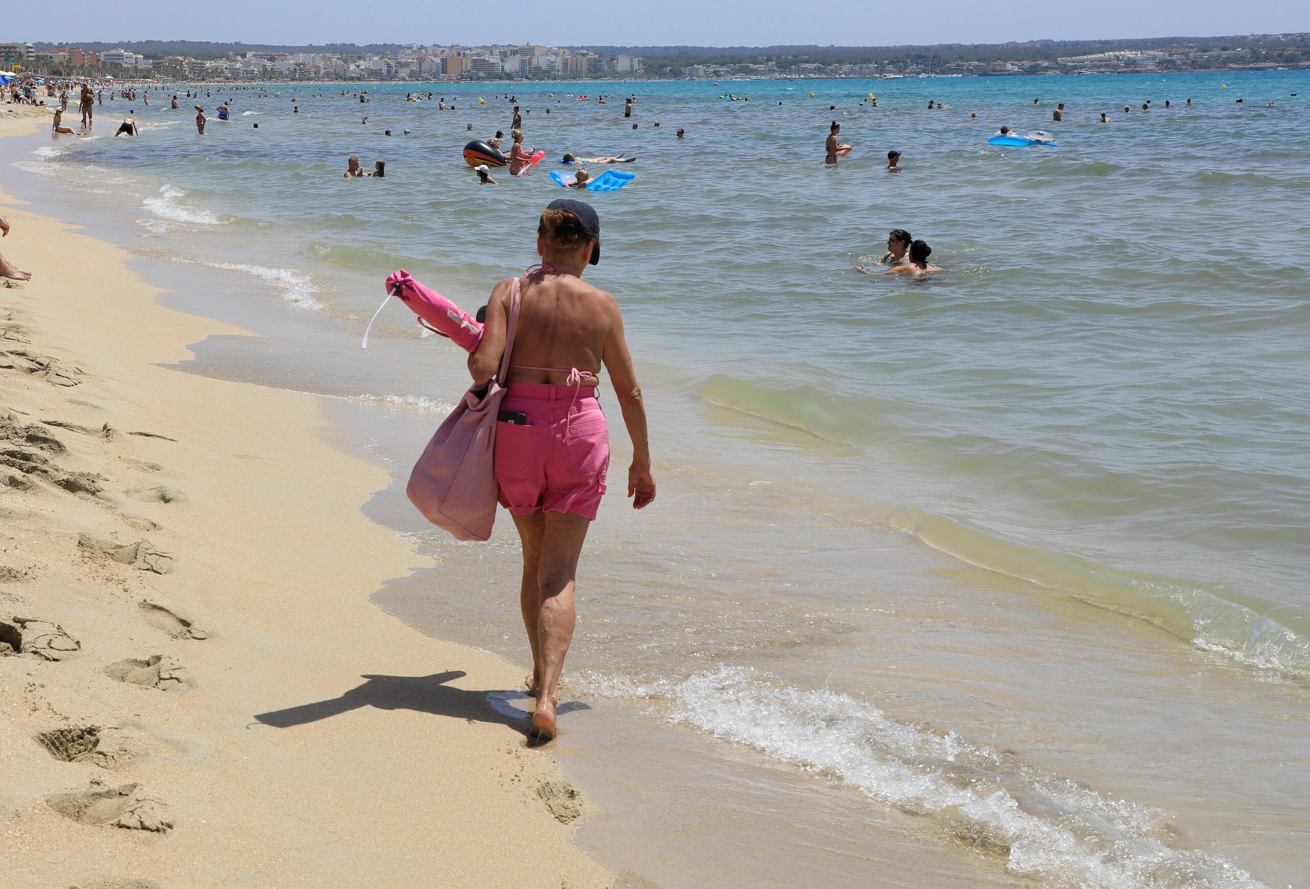 A person in a pink bathing suit walks away from the camera towards the ocean full of people on a sandy beach