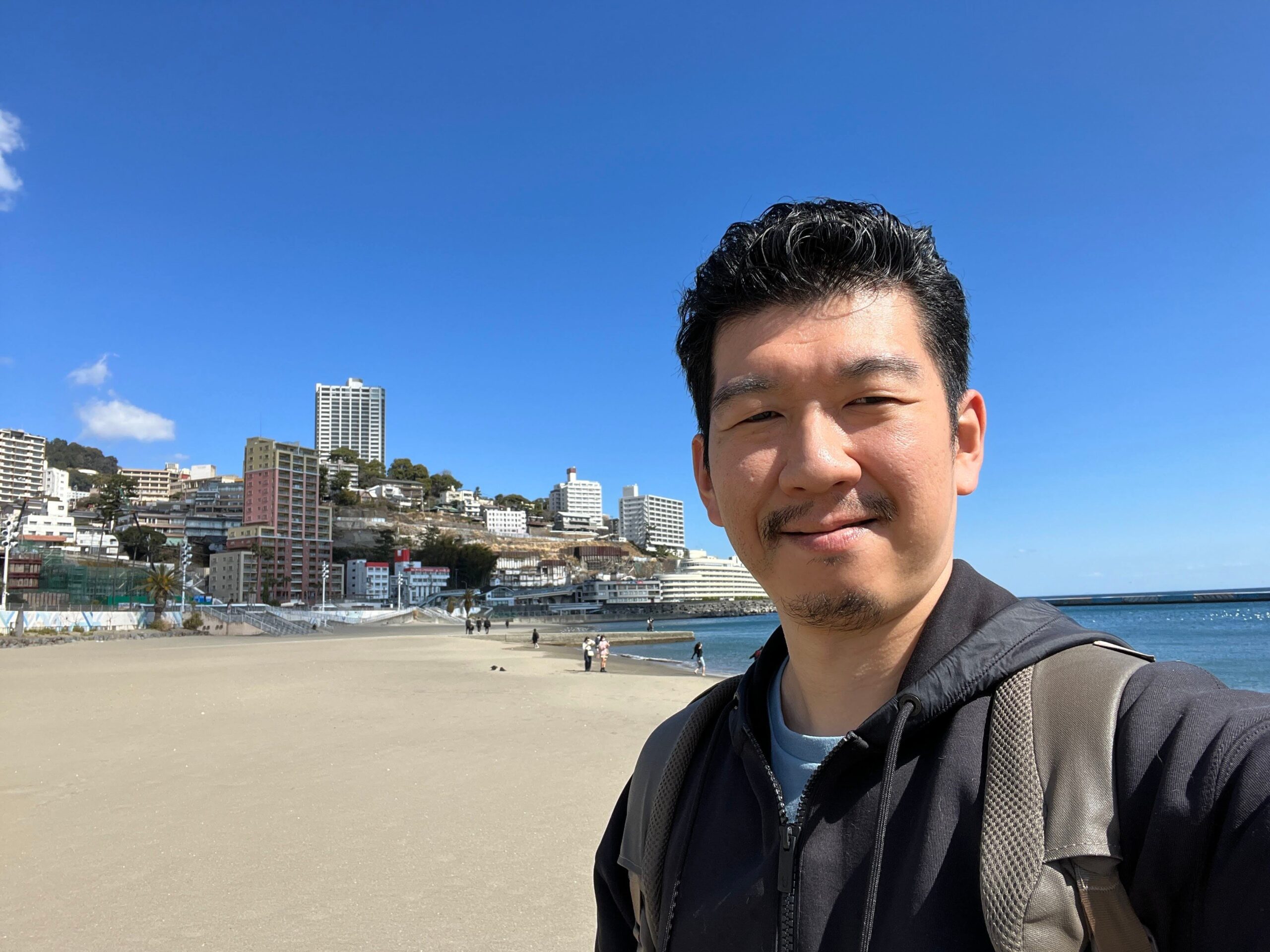 Een man maakt een selfie met het strand van Atami op de achtergrond