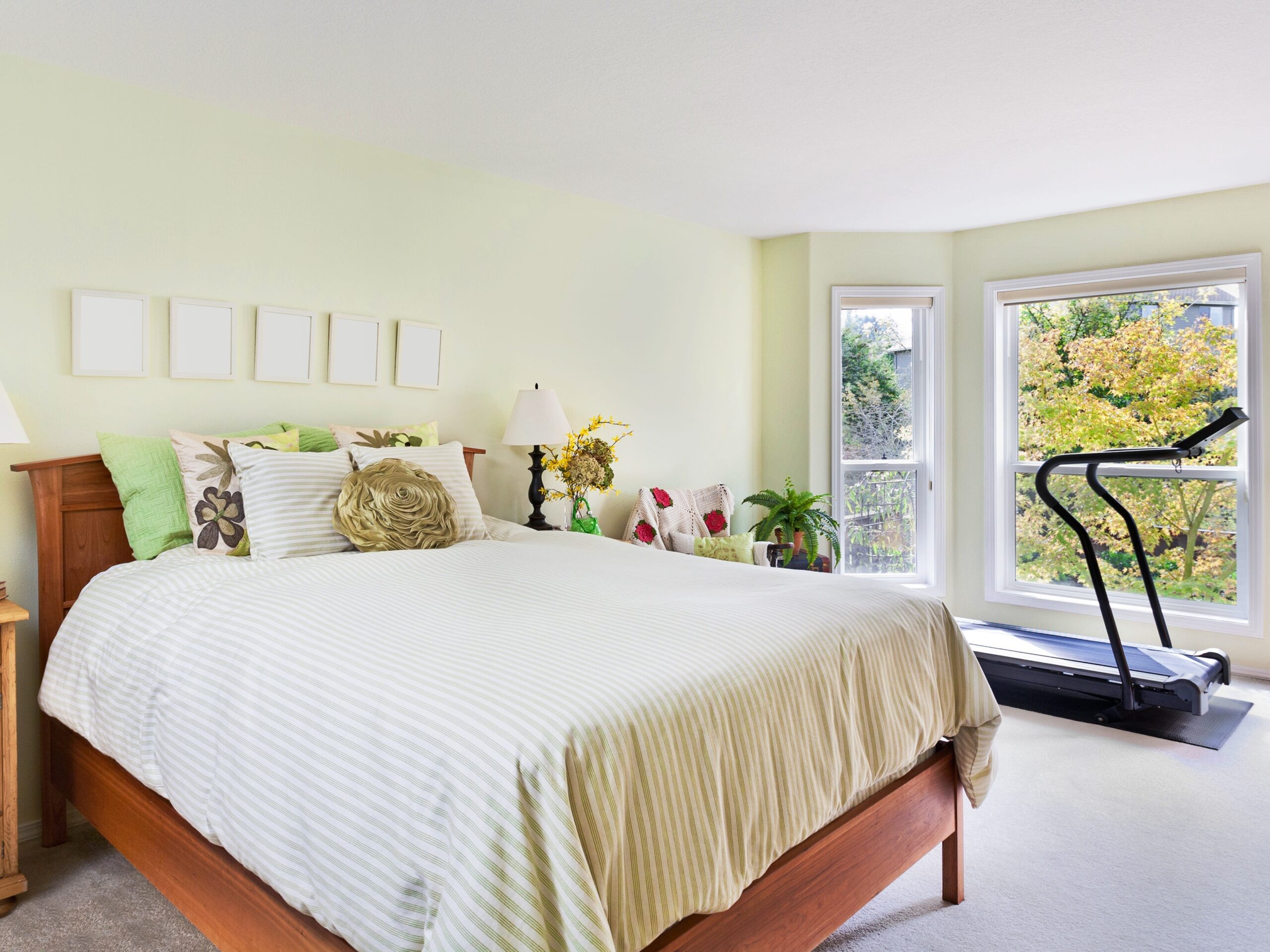 A bedroom with a bed, two nightstands, decor, and a treadmill by the windows.