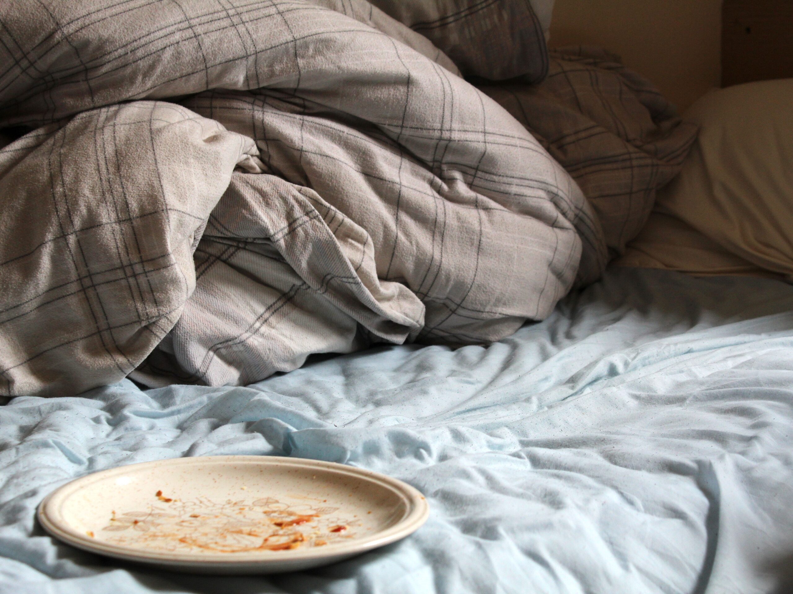 A dirty dish on an unmade bed with a blue sheet and gray comforter and pillows.