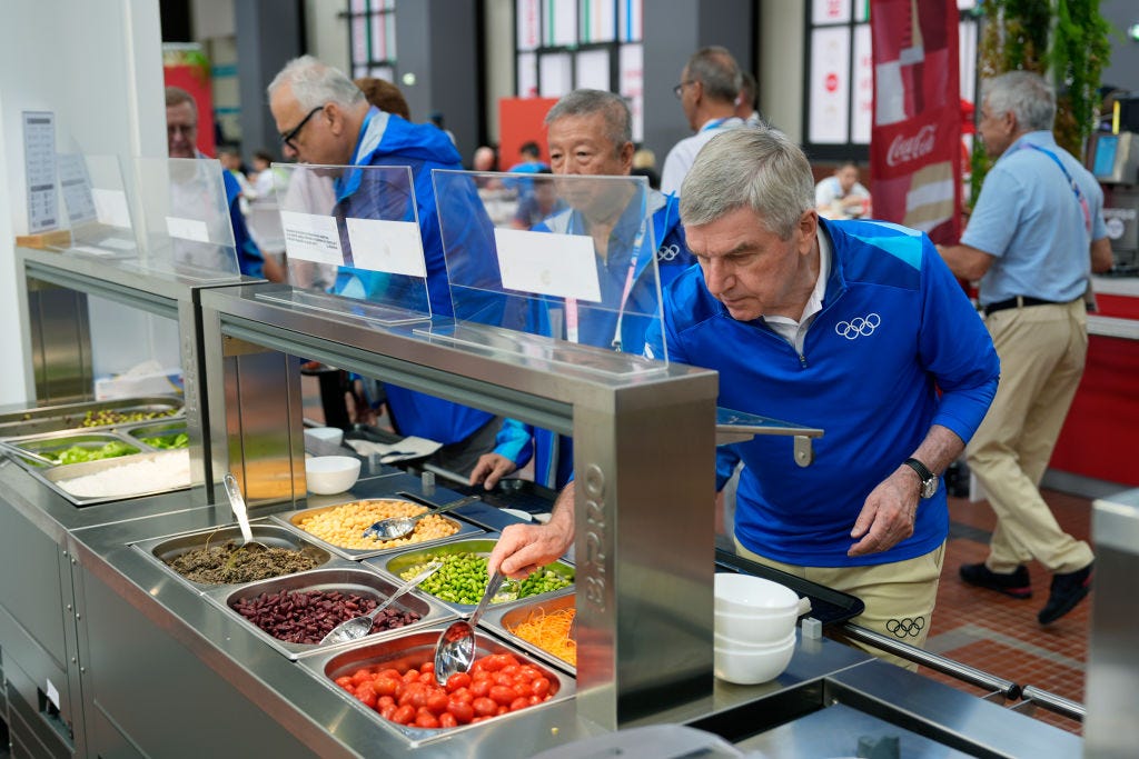 IOC President Thomas Bach schept op uit de saladebar tijdens zijn tour door het Olympisch dorp voorafgaand aan de Olympische Spelen.