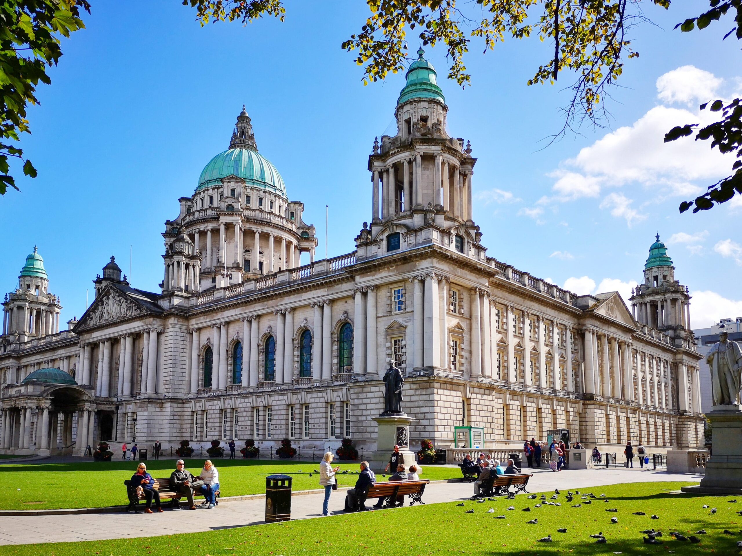 Het gebouw van het stadhuis van Belfast achter een park met bankjes en gras