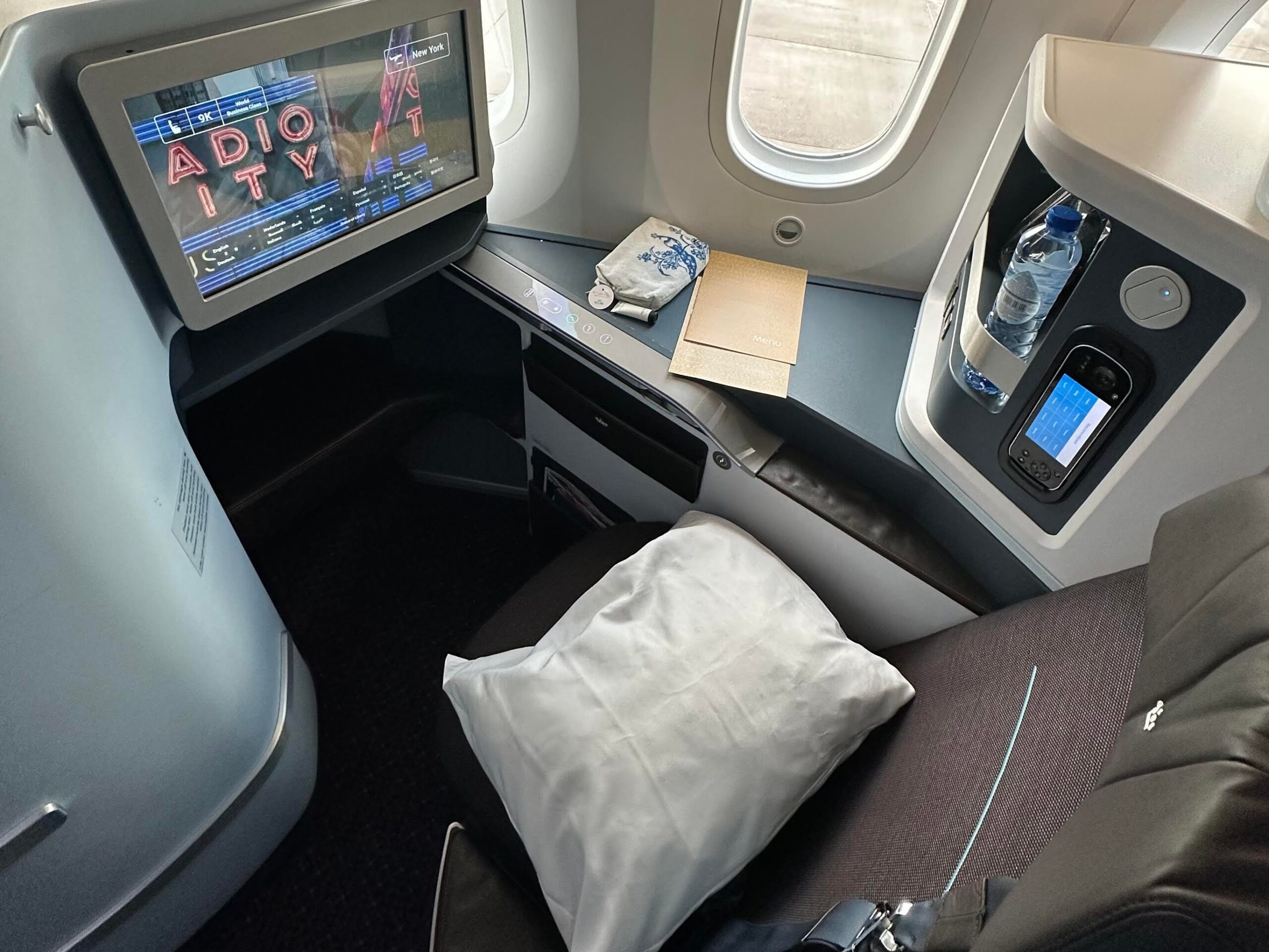 Een business-class stoel met een kussen, een waterfles, een afstandsbediening en een tv met een afbeelding van Radio City Music Hall op het scherm.