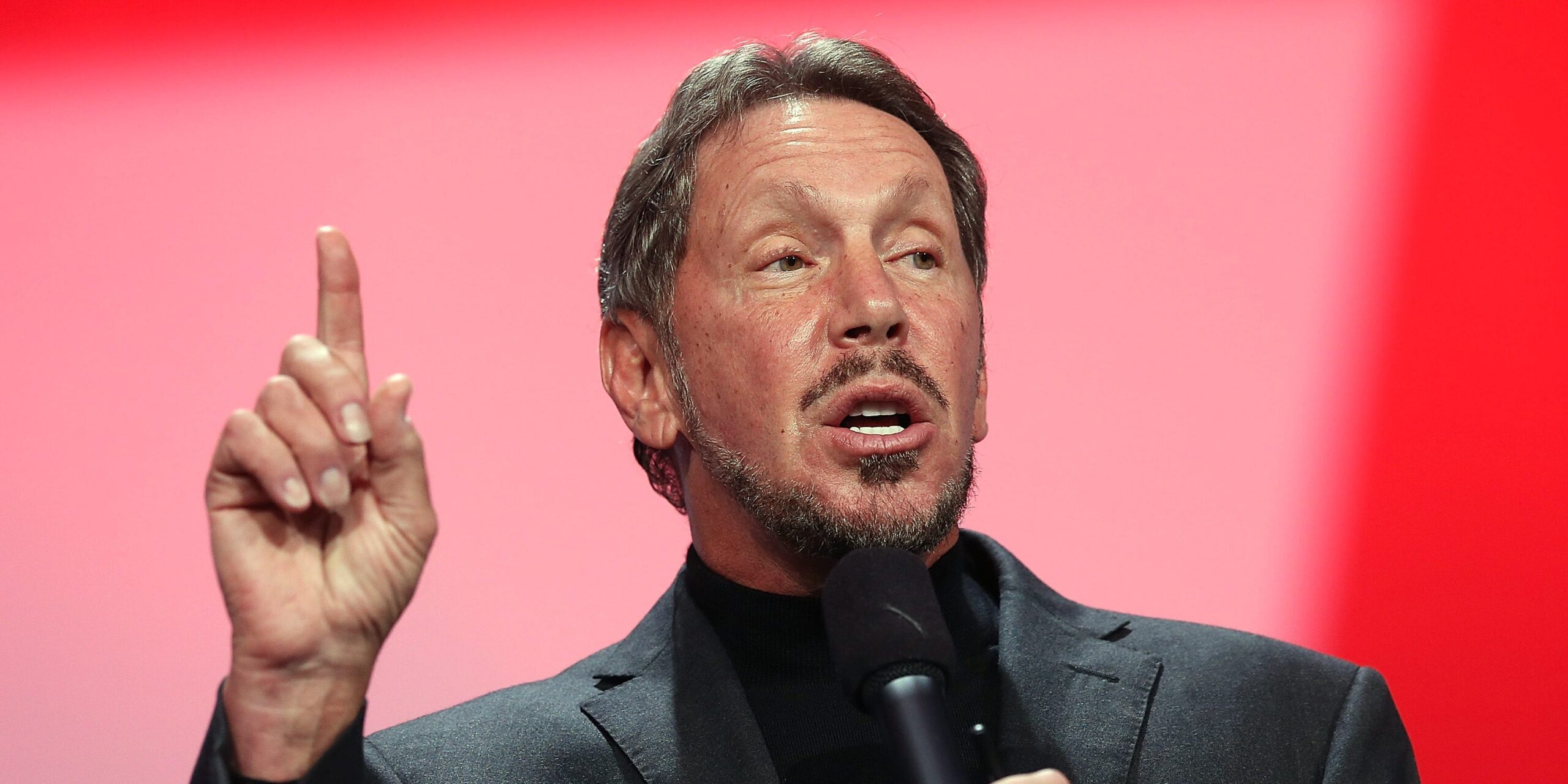 Larry Ellison speaking into a microphone and pointing upward.