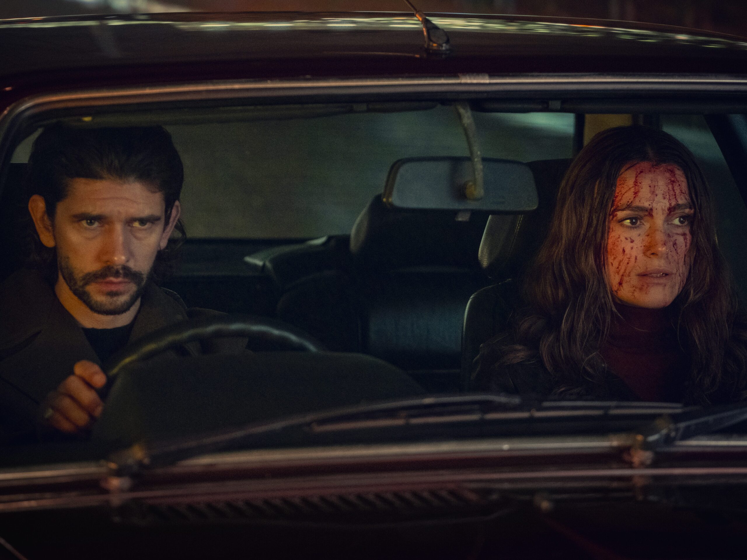 An image of a man with long black hair and a beard sitting in the driving seat of a car. In the passenger seat is a woman with long brown hair whose face is covered in blood.