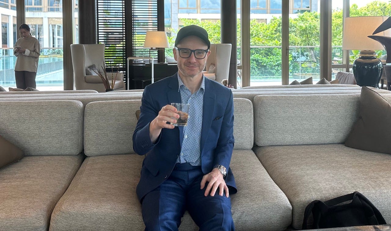 Author Paul Oswell in a suit sitting with a cocktail on a large couch in front of floor-to-ceiling-windows