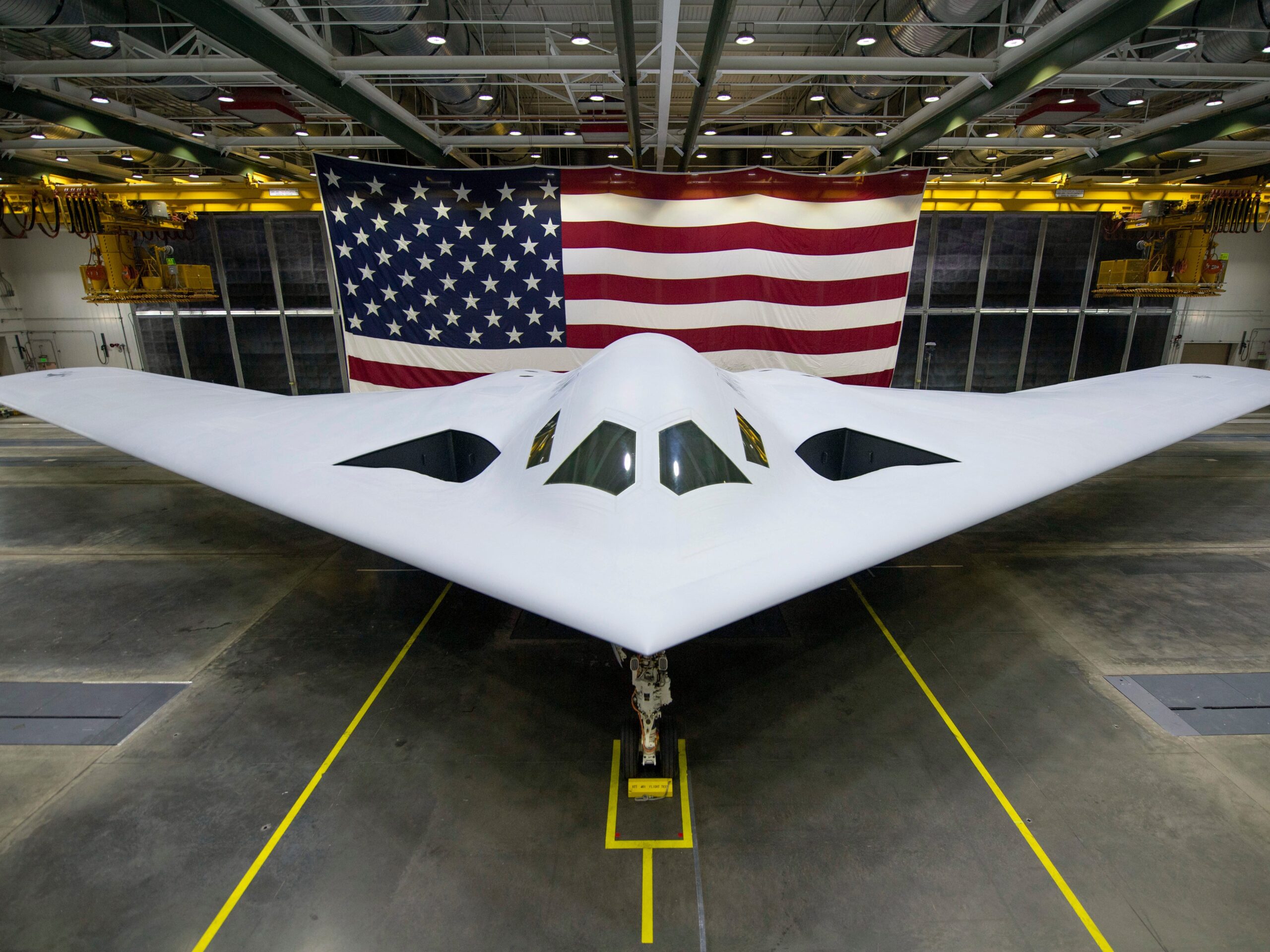 The B-21 Raider is seen in an indoor facility with an American flag behind it.