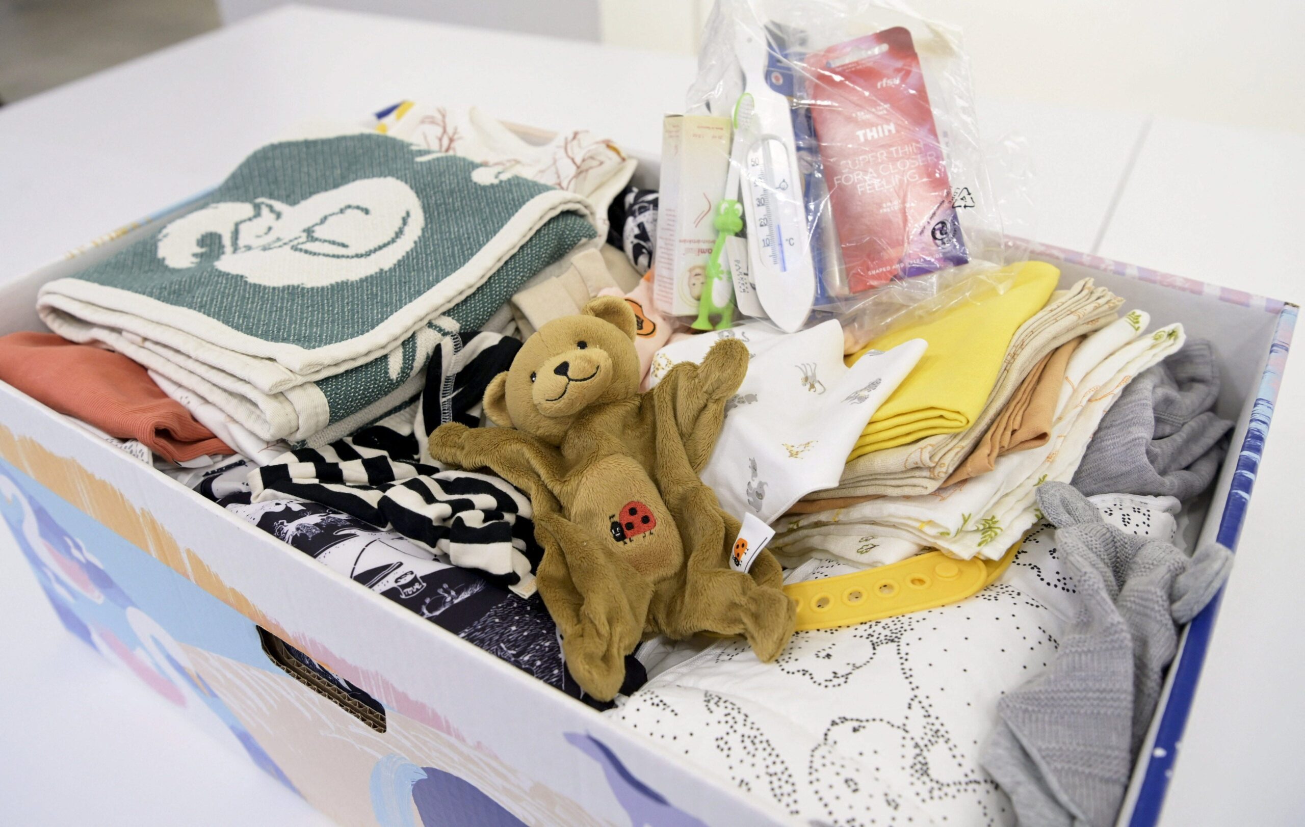 A teddy bear with a ladybug on its stomach rests on clothing, blankets, and other necessities in a Finnish baby box